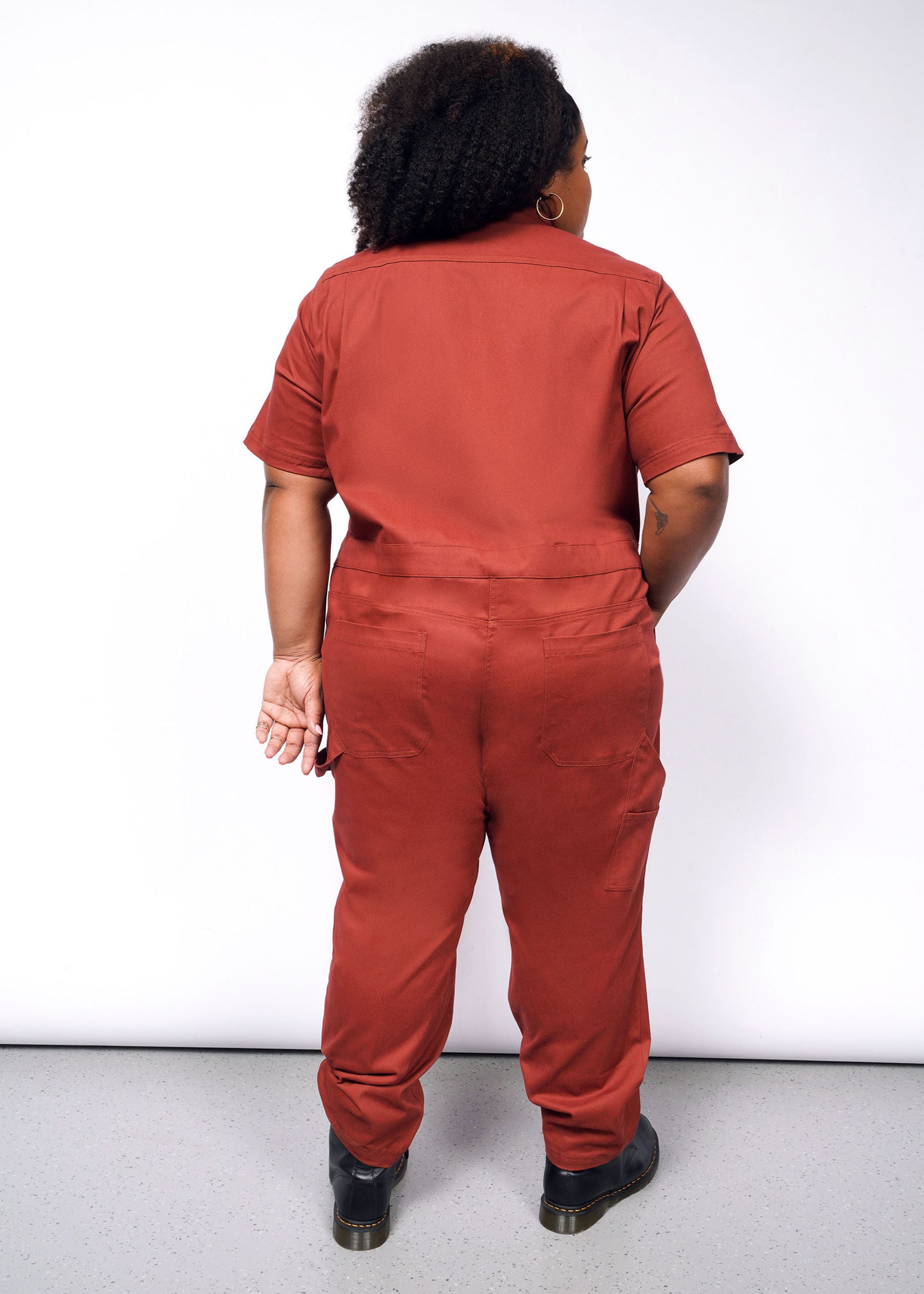 Person with curly hair in The Essential Coverall, a relaxed-fit red jumpsuit made of stretch cotton, and black shoes. They are standing with their back to the camera against a plain light background.