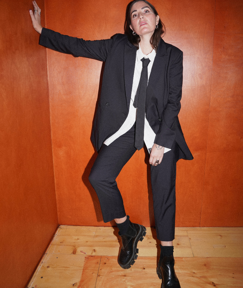 Person with long brown hair leans against wooden wall wearing black blazer, white button up, black tie, black trousers, and black chunky boots. Background is wooden wall with lighter wooden floor.