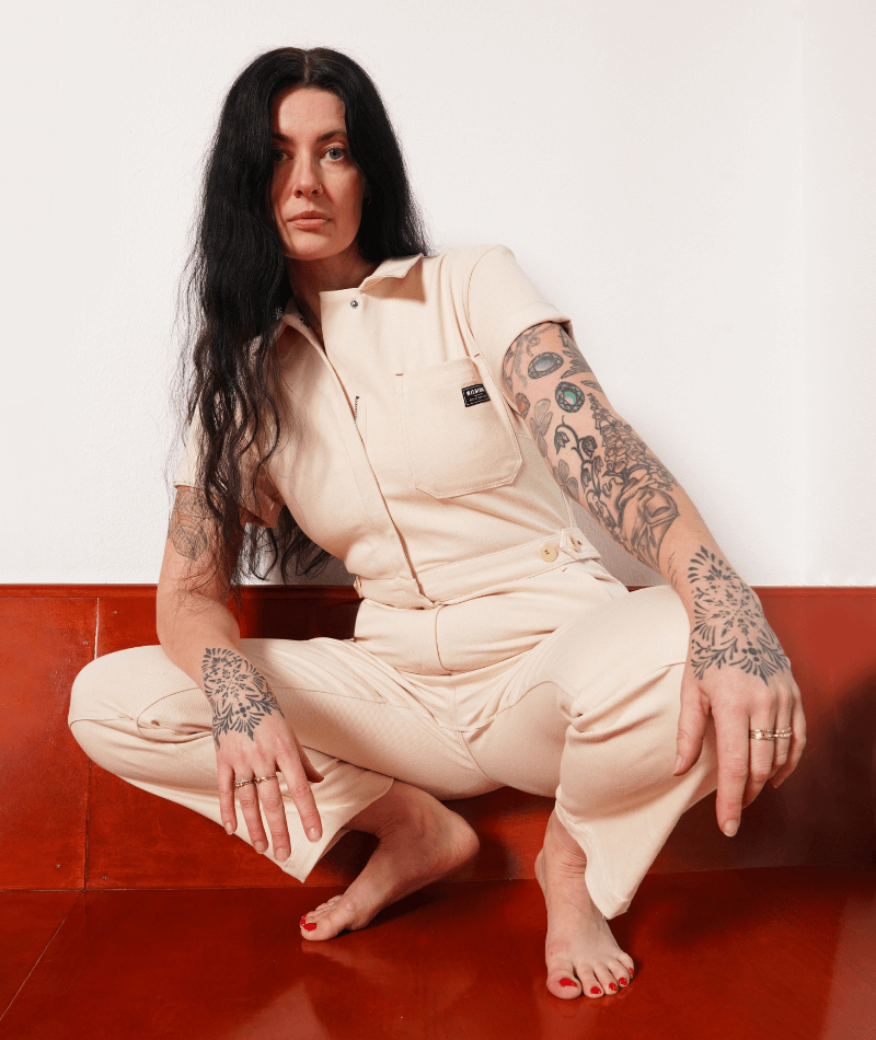 A person with tattoos wearing The Essential Denim High Waisted Coverall crouches on wood paneled platform.