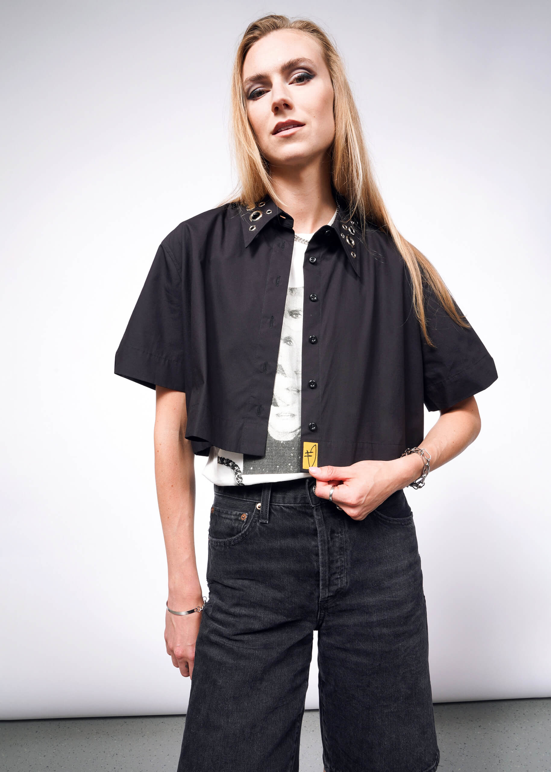 A person with long blond hair in a black cropped jacket and the oversized Debbie Harry X Wildfang Cropped Button Up shirt stands confidently against a plain white background.