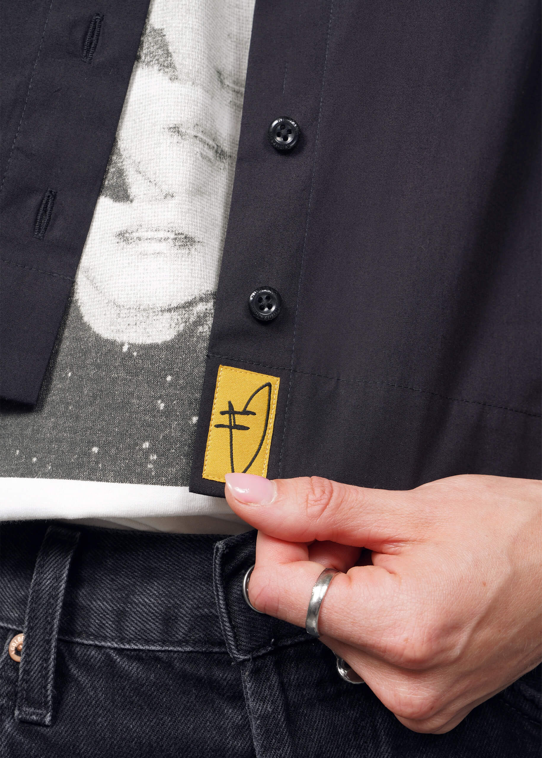 A person holds the hem of a black shirt, revealing a yellow logo tag with a black symbol. Below, a white t-shirt from the Debbie Harry X Wildfang collection is partially visible. They wear black jeans and a silver ring.