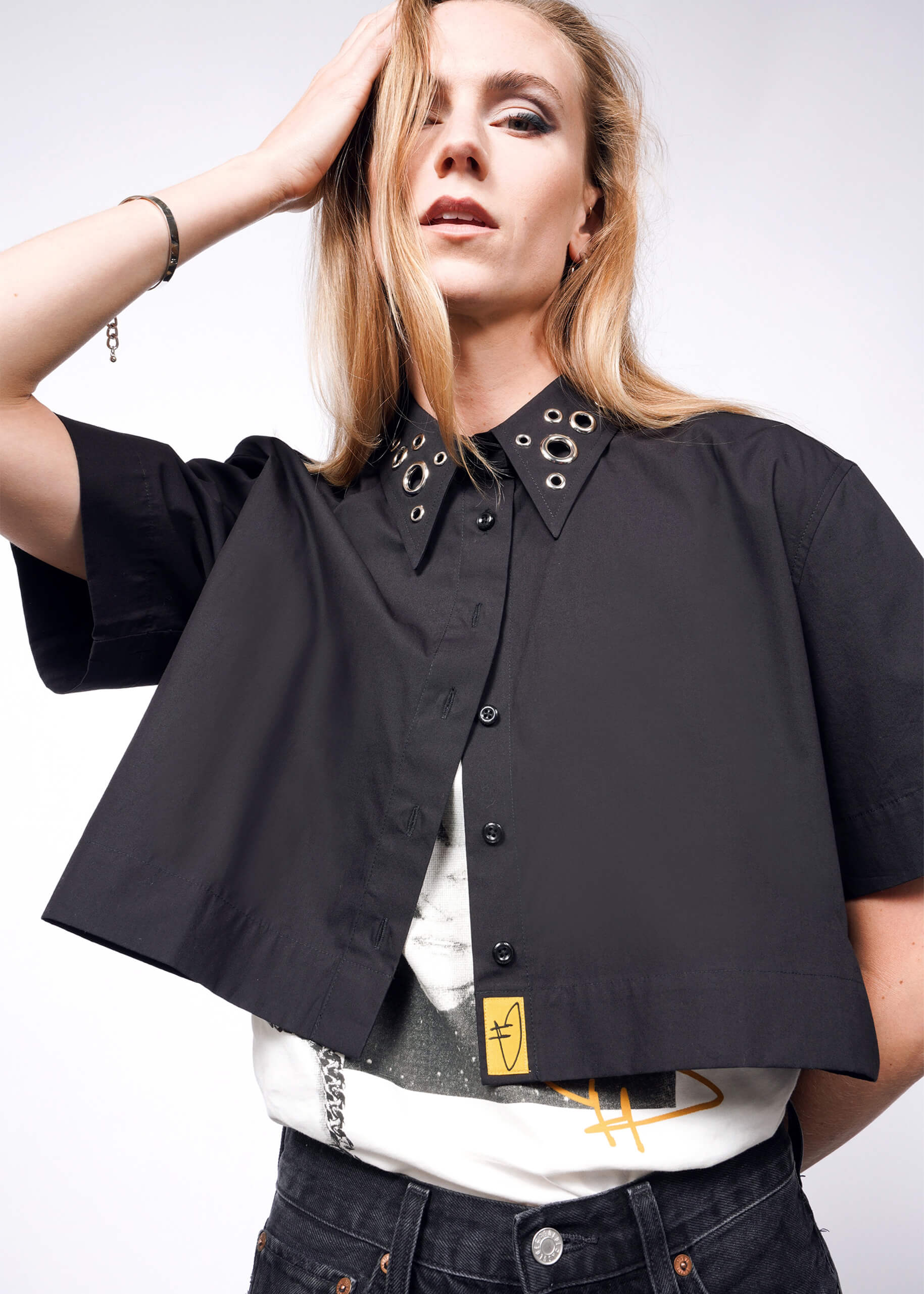 A person with long blonde hair poses wearing a short-sleeved, black Debbie Harry X Wildfang Cropped Button Up featuring a grommet-adorned collar. They sport an oversized fit printed T-shirt and dark jeans against a plain white background, their mouth slightly open.