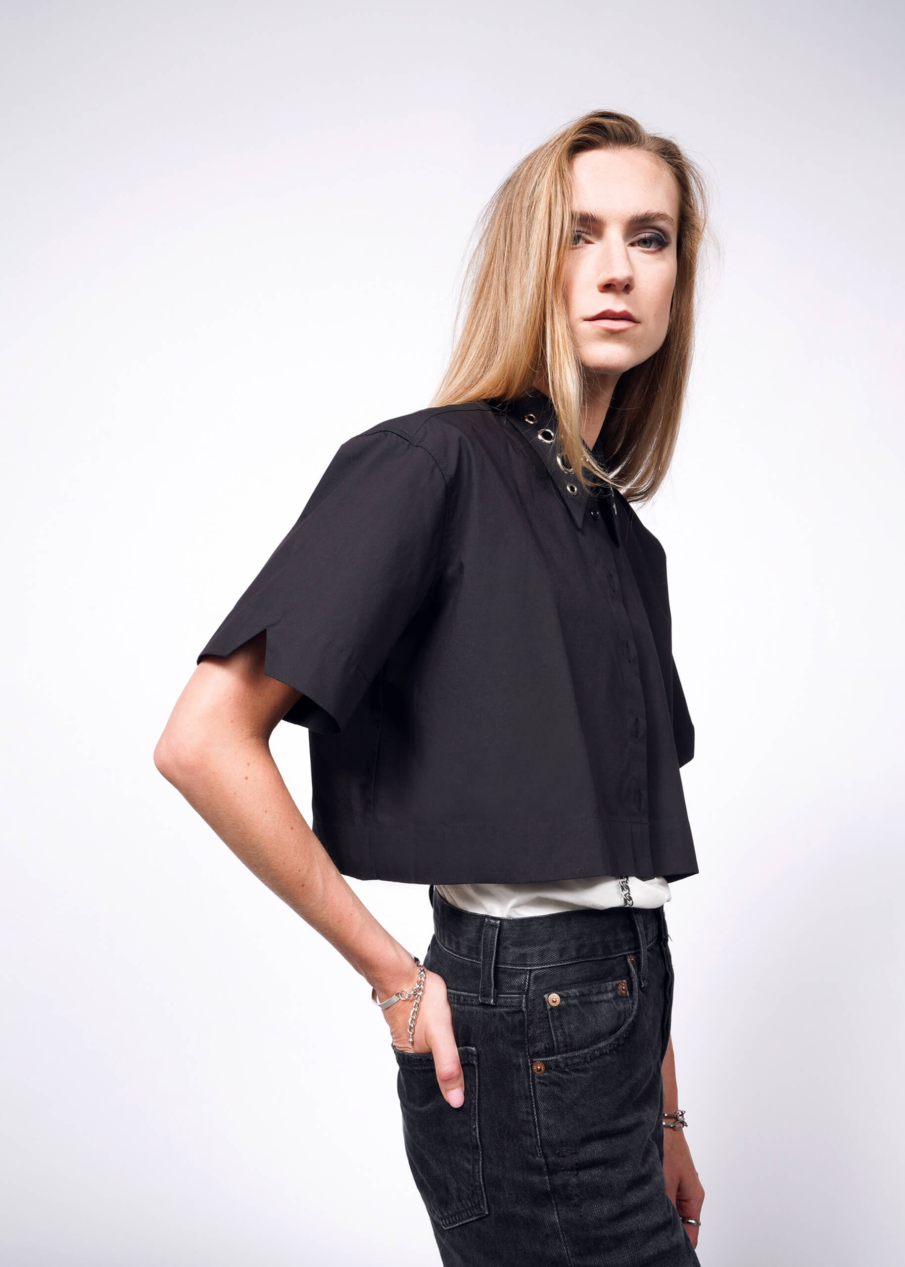 A person with long, light brown hair is wearing a black Debbie Harry X Wildfang Cropped Button Up and dark jeans. They stand against a plain white background, maintaining a neutral expression while looking directly at the camera.