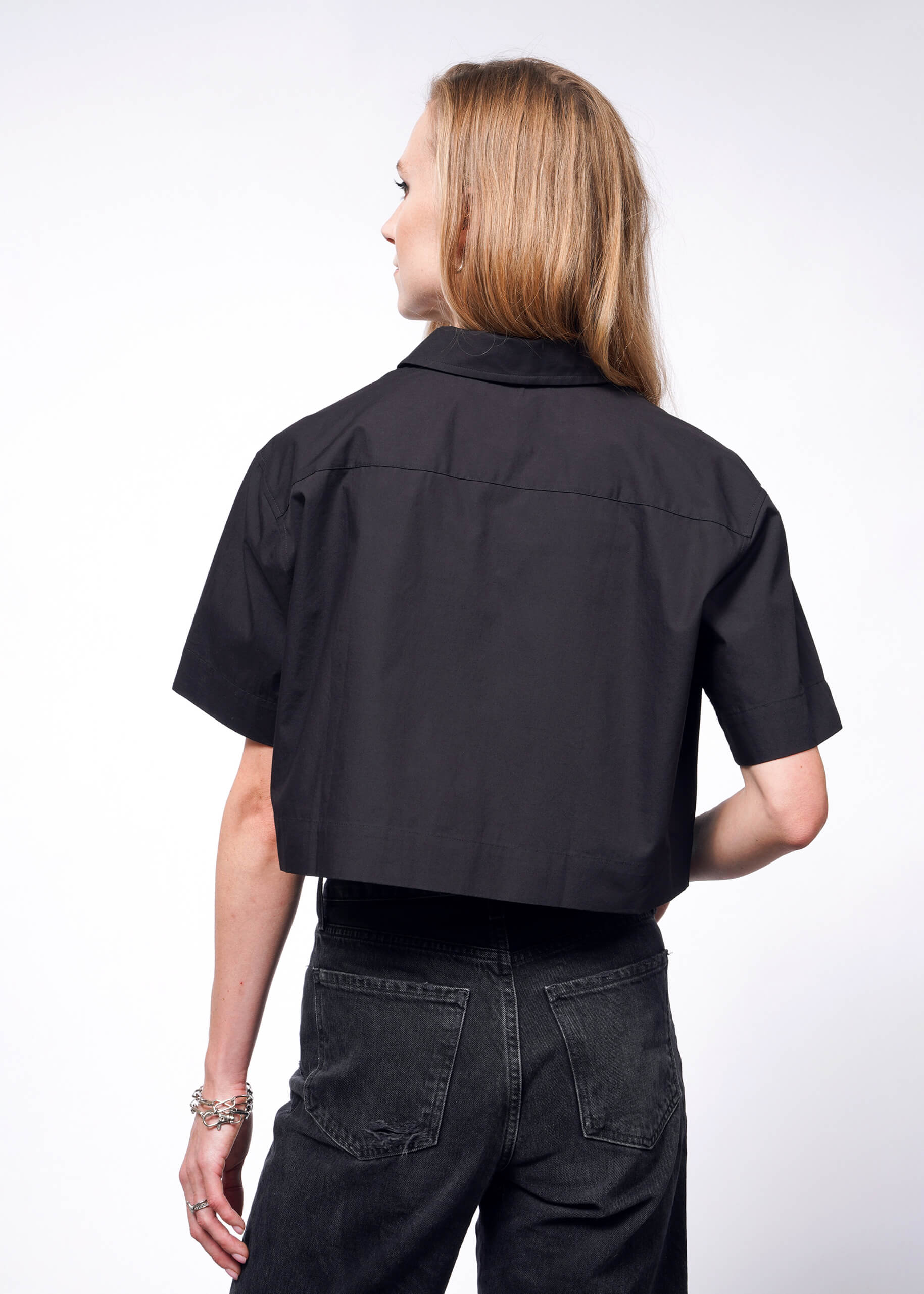 A person with long hair, seen from the back against a white background, is wearing an oversized fit, black Debbie Harry X Wildfang Cropped Button Up and dark jeans.