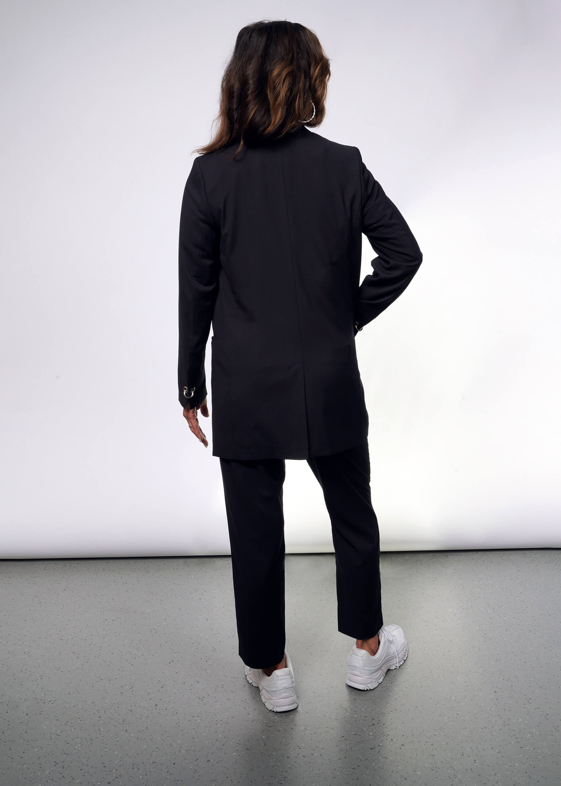 A person with long, wavy hair faces away from the camera, donning a sharp Debbie Harry X Wildfang Double Breasted Blazer and sleek white sneakers. The look is framed by a plain white wall and grey floor, subtly enhanced by metal detailing in the ensemble.