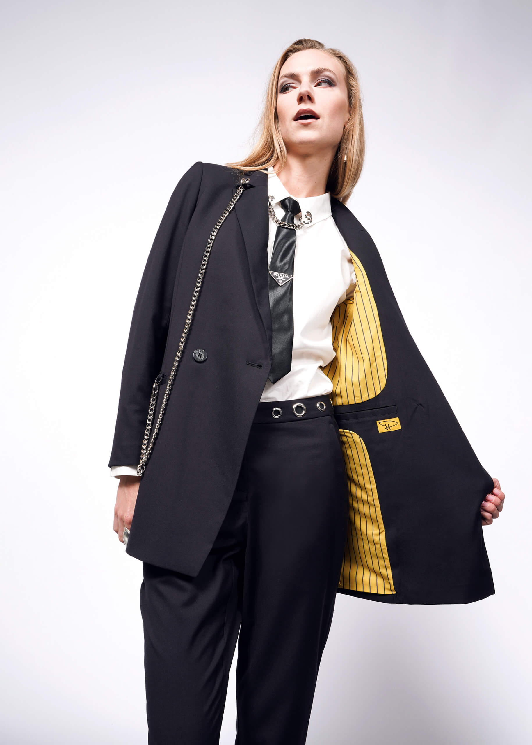 Dressed in a stylish black suit featuring the Debbie Harry X Wildfang Slim Crop Pant, a person poses confidently. They reveal the blazers yellow-striped lining over a white shirt and black tie, accentuated by chain details, embodying a punk aesthetic against a plain white background.