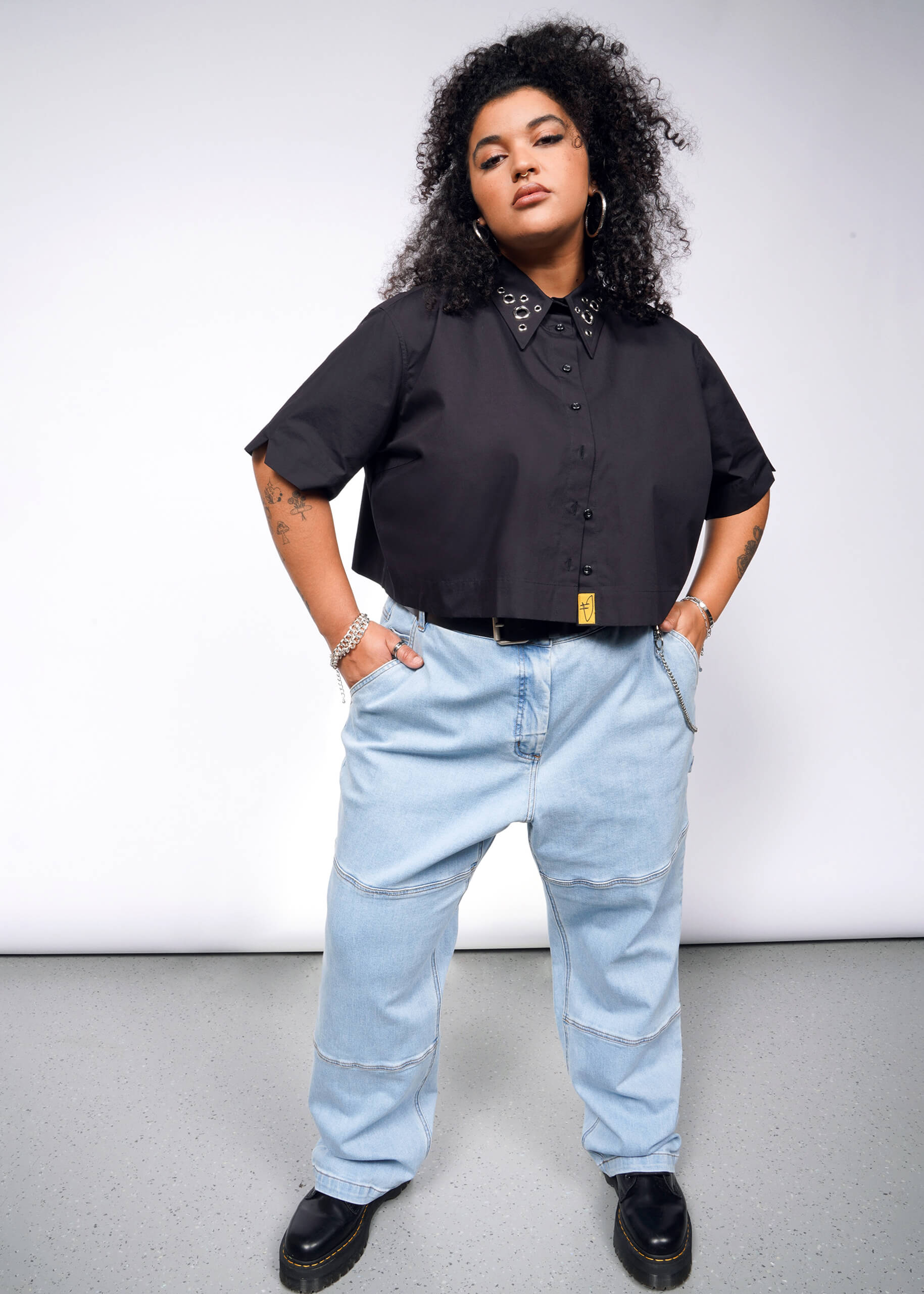 A person with curly hair confidently stands in an oversized Debbie Harry X Wildfang Cropped Button Up, light blue jeans, hoop earrings, a bracelet, and black shoes against a plain backdrop, hands in pockets.