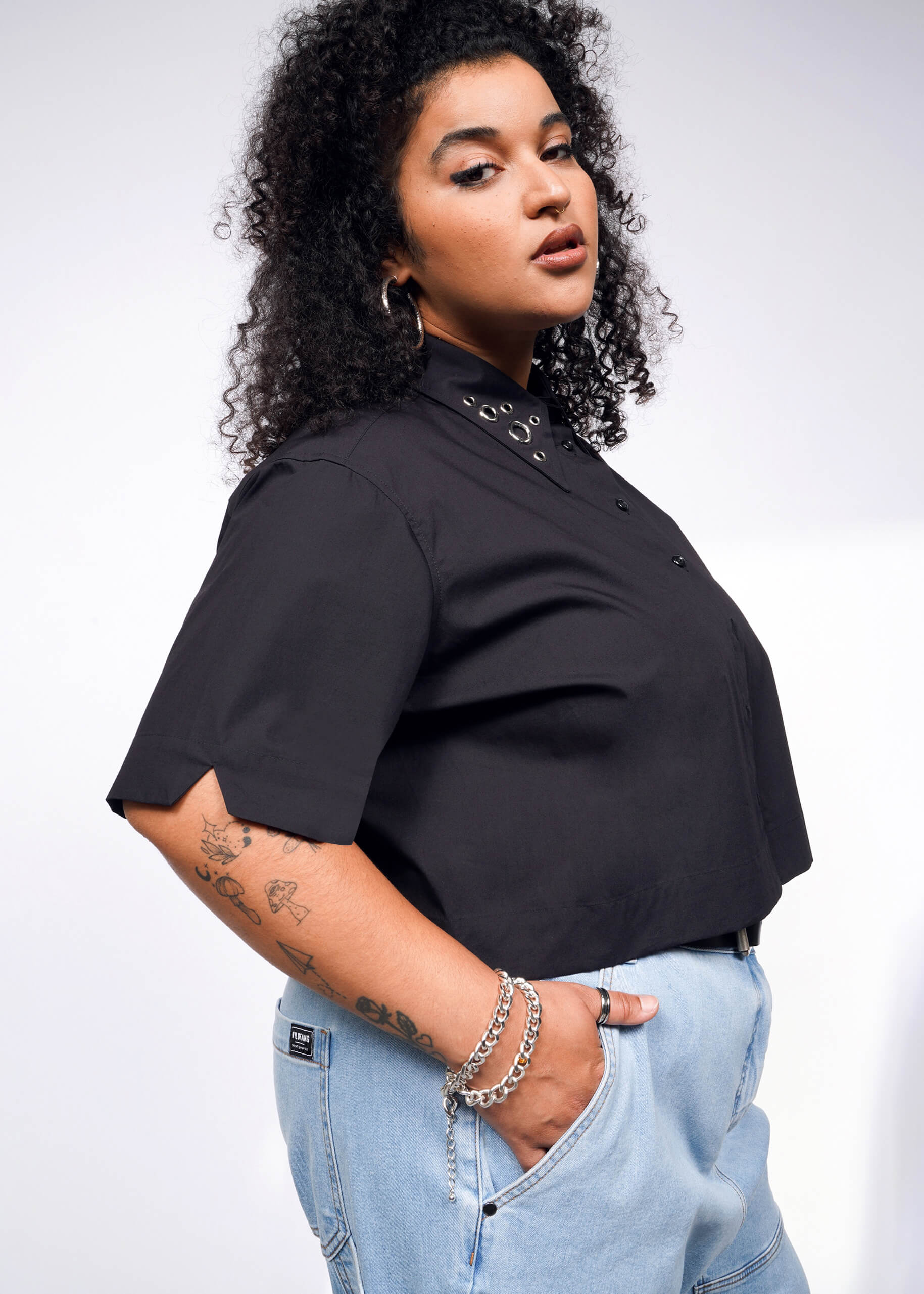 A woman with curly hair wears the Debbie Harry X Wildfang Cropped Button Up and light blue jeans, flaunting hoop earrings and bracelets. Her forearm tattoos show as she stands against a light background, hinting at Debbie Harrys capsule style while glancing over her shoulder.