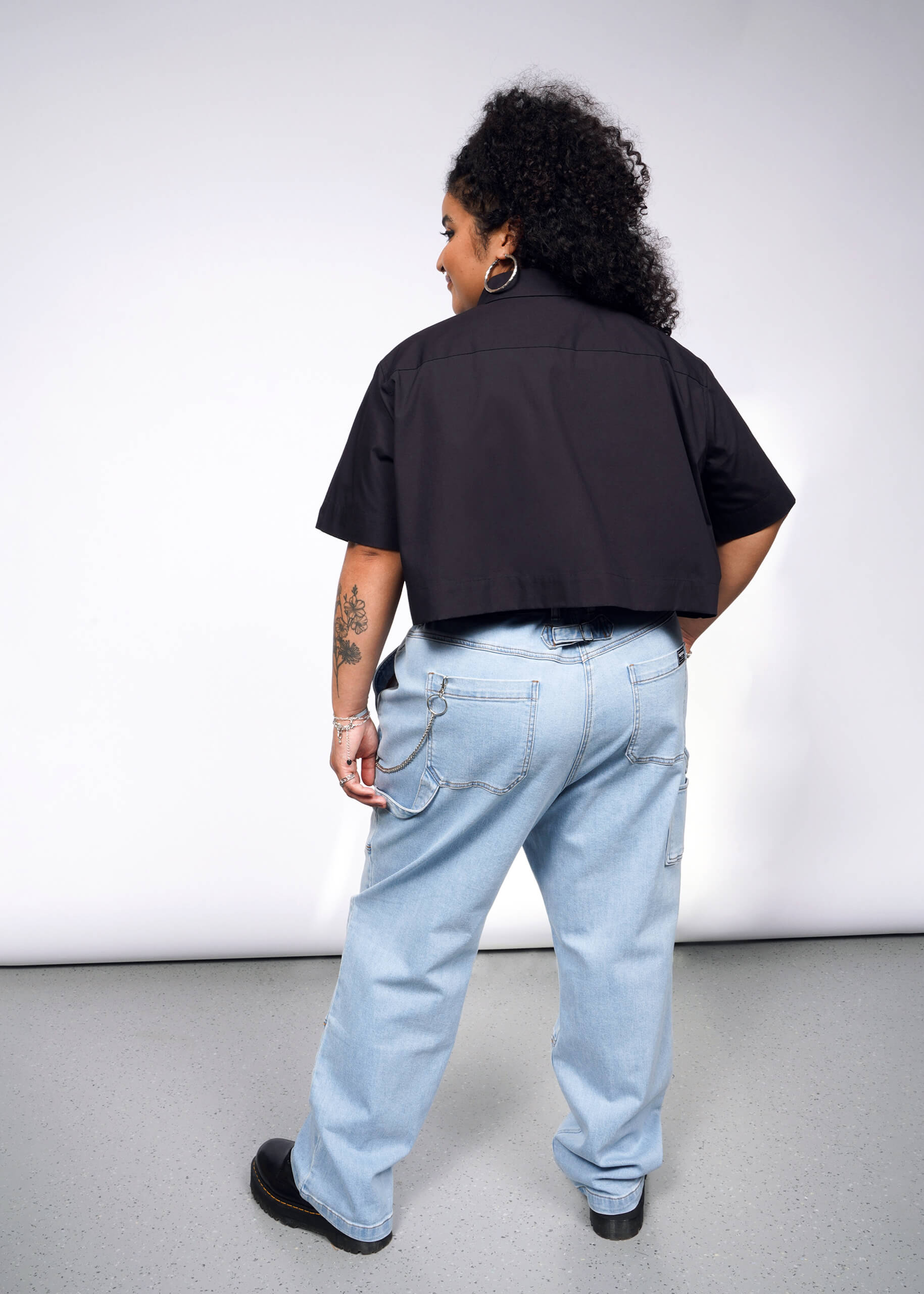 A person with curly hair, wearing the Debbie Harry X Wildfang Cropped Button Up in black and light blue jeans with chain details, stands facing away in a room with a light gray floor and wall. They have visible tattoos on their left arm.