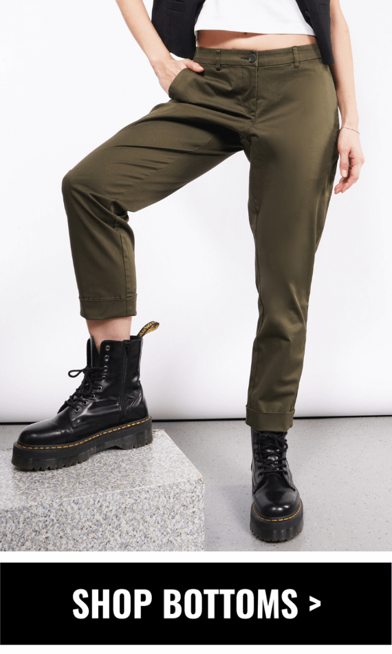 Shows model from the waist down wearing The Essential Trouser in Spruce color and Doc Martens with one foot up on marble block. Button underneath image says SHOP BOTTOMS.