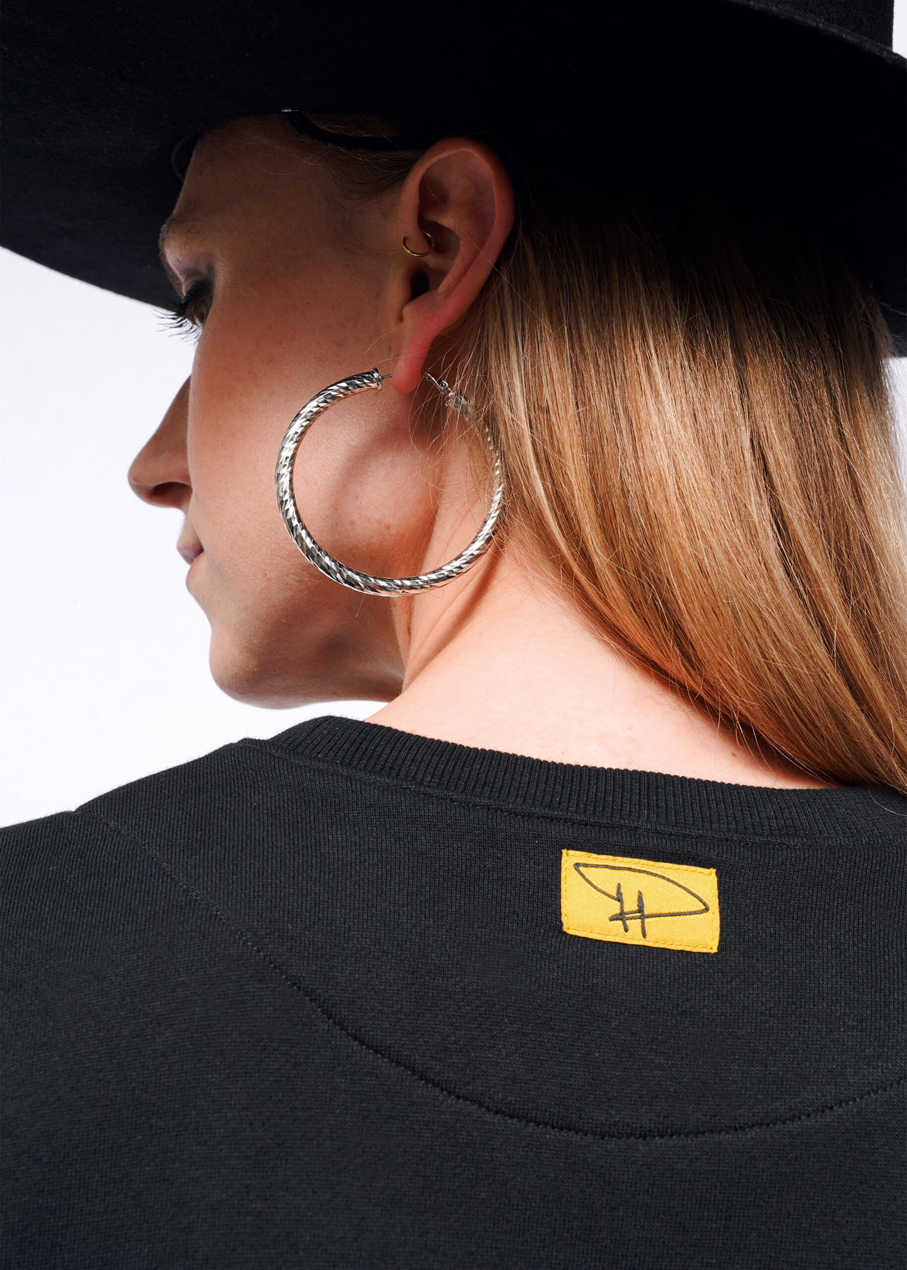 A person with long hair, a large black hat, and silver hoop earrings faces away from the camera. Their black shirt has a yellow label with a stylized H, and theyre wearing the Debbie Harry X Wildfang Convertible Sweatshirt, featuring chic removable sleeves.