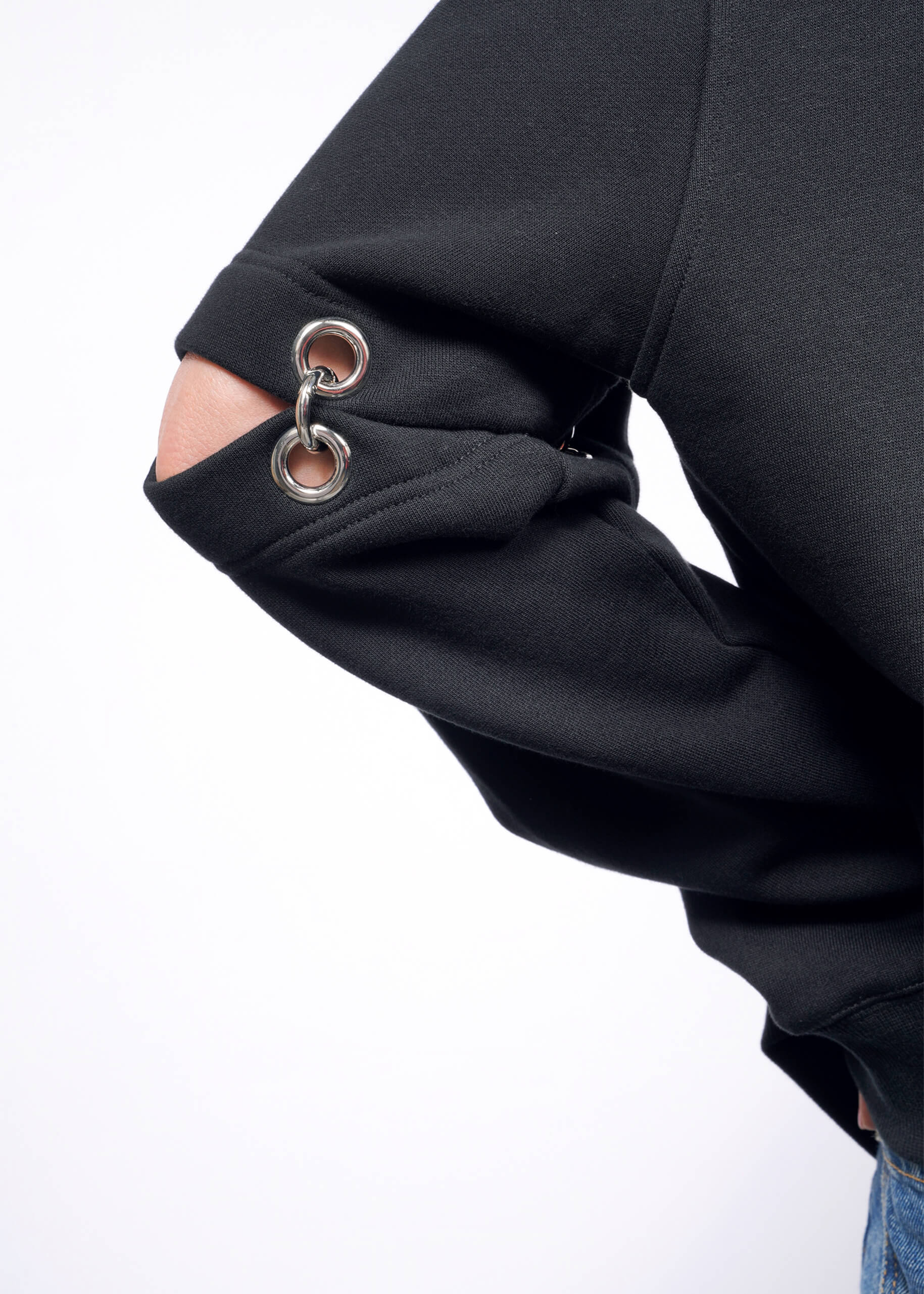 A person showcases the Debbie Harry X Wildfang Convertible Sweatshirt, a black jacket with unique removable sleeve cut-outs featuring metal grommets, as their hand rests on their hip against a white background.