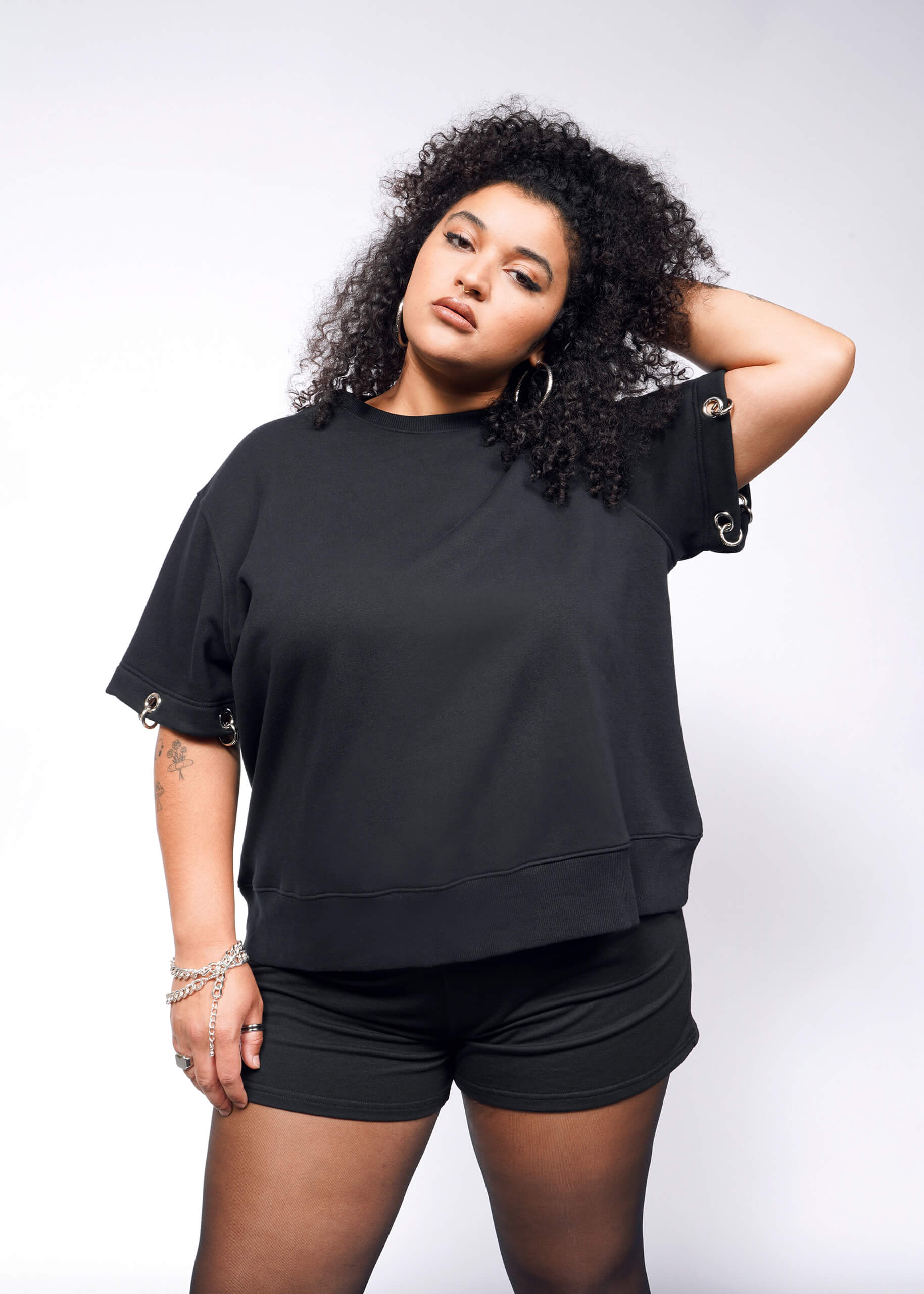 A person with curly hair confidently poses, one hand behind their head, wearing the black Debbie Harry X Wildfang Convertible Sweatshirt with metal ring details and matching shorts. Their other hand rests by their side adorned with bracelets against a plain white background reflecting an edgy style.