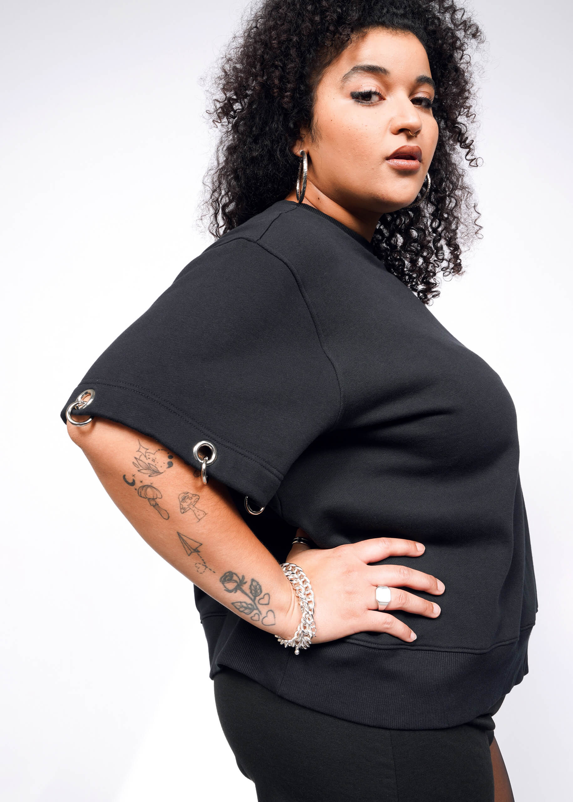 Against a white background, a person with curly hair showcases their confidence. Theyre wearing hoop earrings and the Debbie Harry X Wildfang Convertible Sweatshirt in black with removable sleeves featuring metal eyelets. Tattoos adorn one arm, complemented by a chain bracelet and ring.