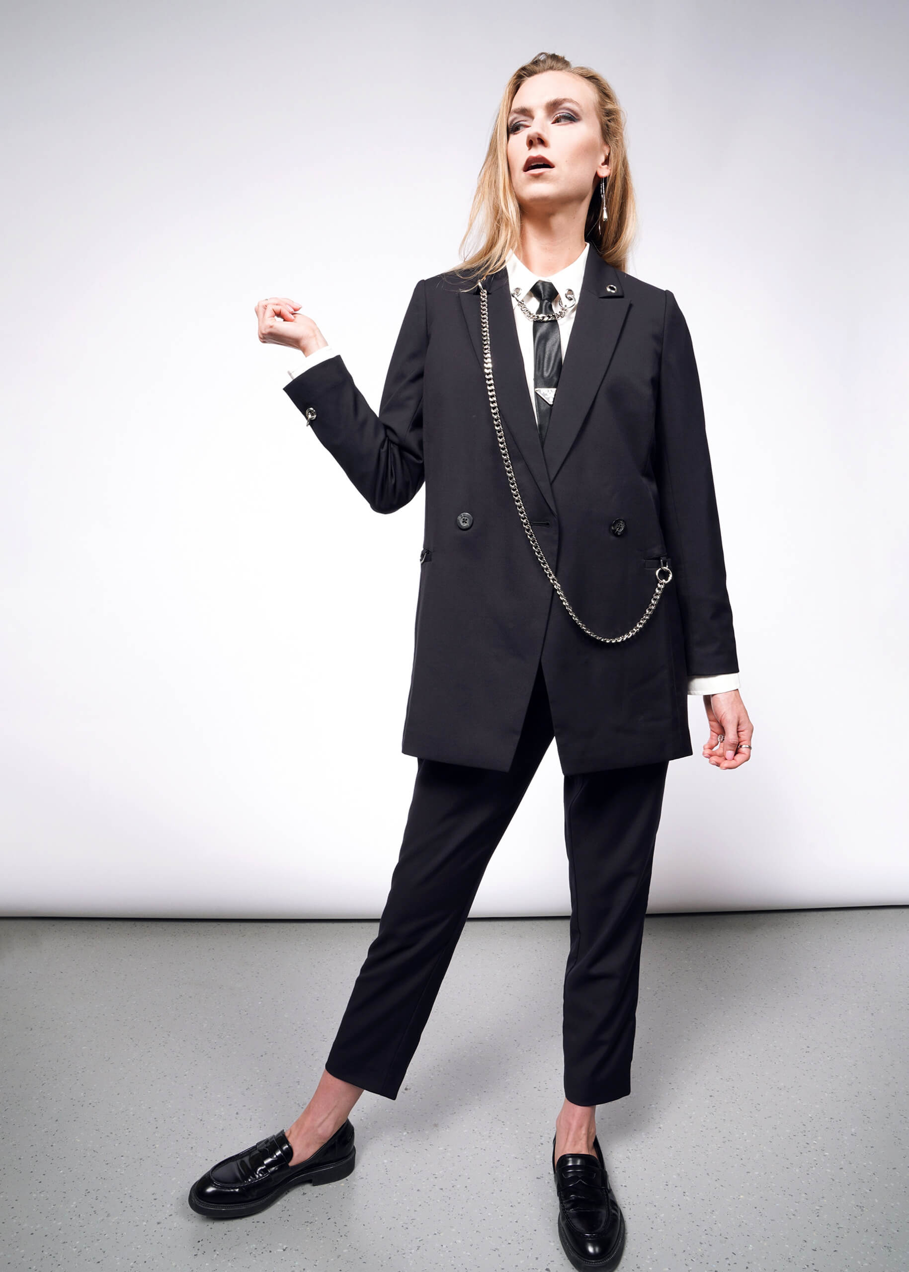 Confidently posing against a plain backdrop, she embodies elegance in a black tailored suit from the Wildfang collection. Her white shirt complements the Debbie Harry X Wildfang Double Breasted Blazers chain accessory and metal detailing, while black loafers complete her bold, sophisticated look.