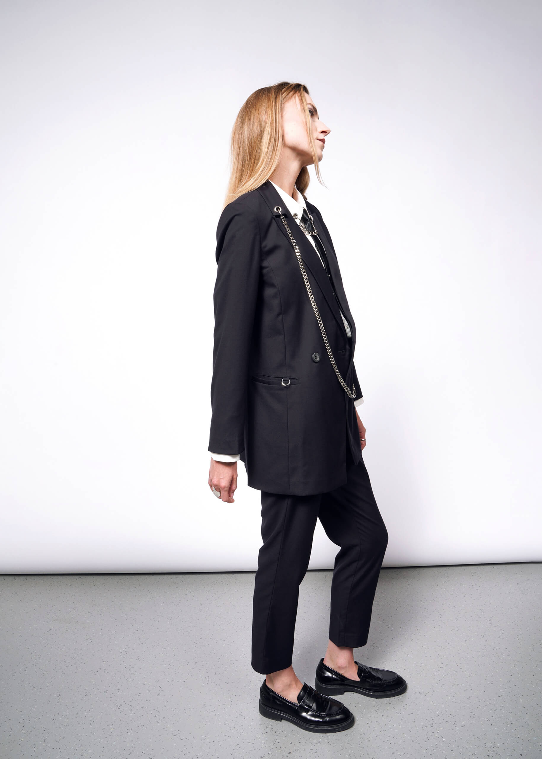 A person with long hair in profile against a plain background wears a Debbie Harry X Wildfang Double Breasted Blazer with metal detailing. They pair it with shiny black loafers, looking upward, their hands relaxed at their sides.