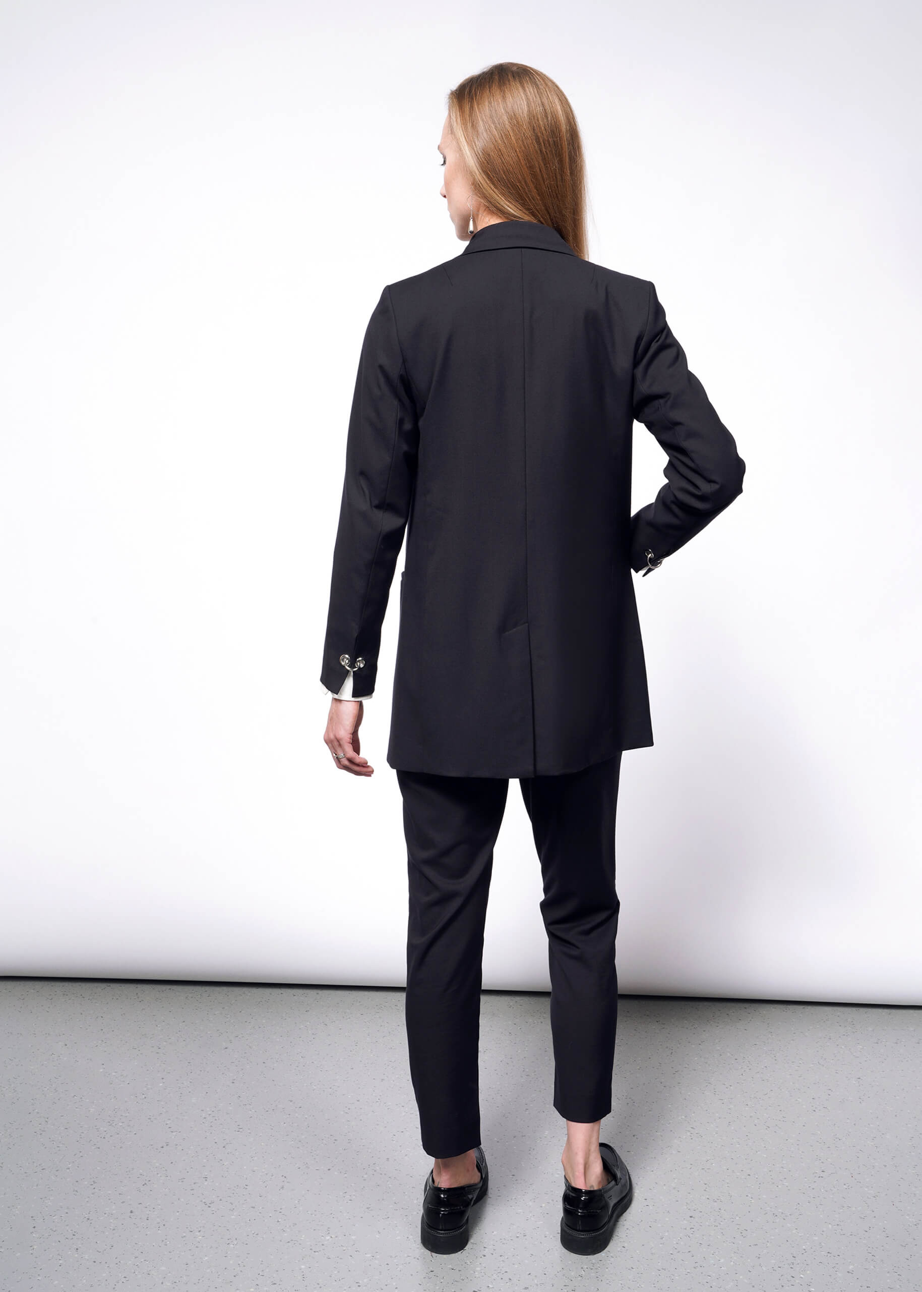 A woman with long hair, facing away on a neutral background, wears a Debbie Harry X Wildfang Double Breasted Blazer with matching pants, complemented by black shoes with metal detailing.