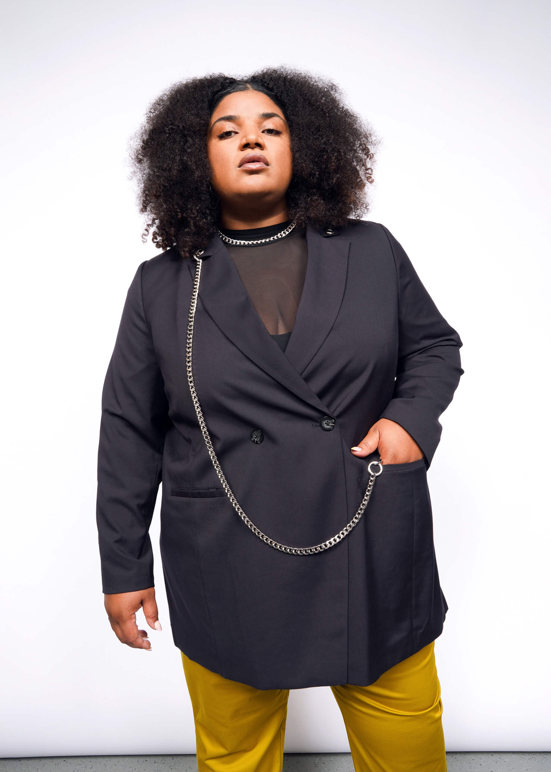 A person with curly hair confidently poses in a stylish Debbie Harry X Wildfang Double Breasted Blazer, featuring intricate metal detailing over a sheer black top and vibrant yellow pants, set against a plain white background.