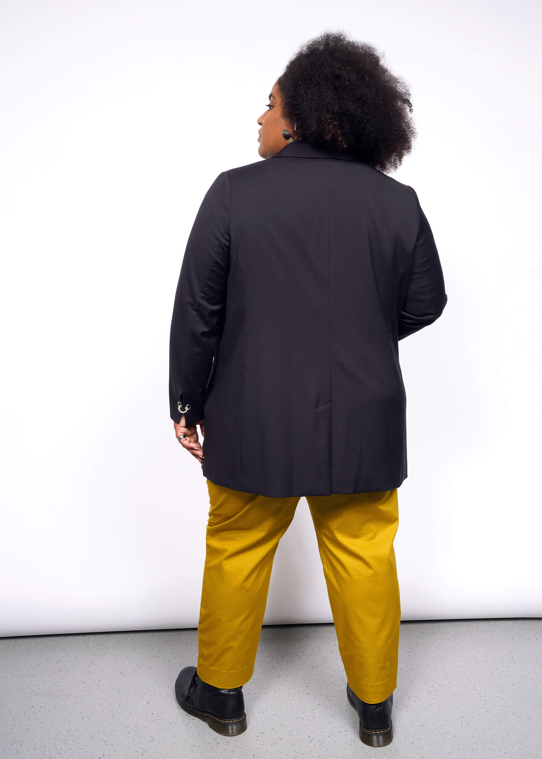 Showcasing a Debbie Harry X Wildfang Double Breasted Blazer with subtle metal details, the person pairs it stylishly with mustard yellow pants and black shoes. Their curly hair cascades down their back as they stand against a pristine white background on a light gray floor.