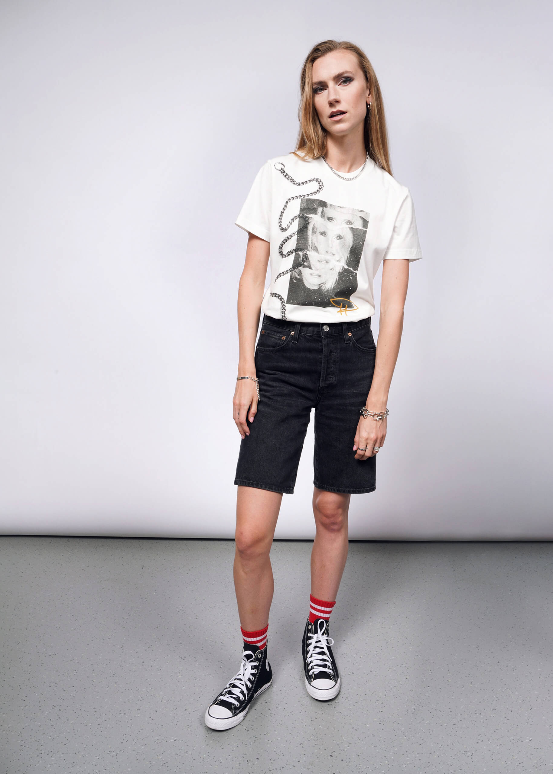 Wearing the Debbie Harry X Wildfang Graphic Tee with black knee-length shorts, black Converse sneakers, and red striped socks, a long-haired person exudes punk style reminiscent of Debbie Harry. They stand relaxed on a gray speckled floor against a neutral background.