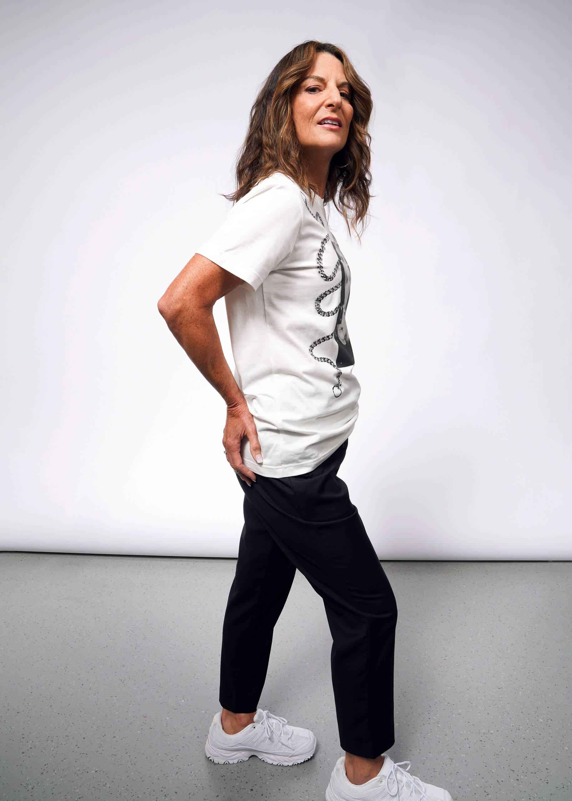 A person with long hair stands confidently in a Debbie Harry X Wildfang Graphic Tee and black pants. Channeling a punk aesthetic, they wear white sneakers, reminiscent of Debbie Harrys iconic style, posed against a plain gray background.