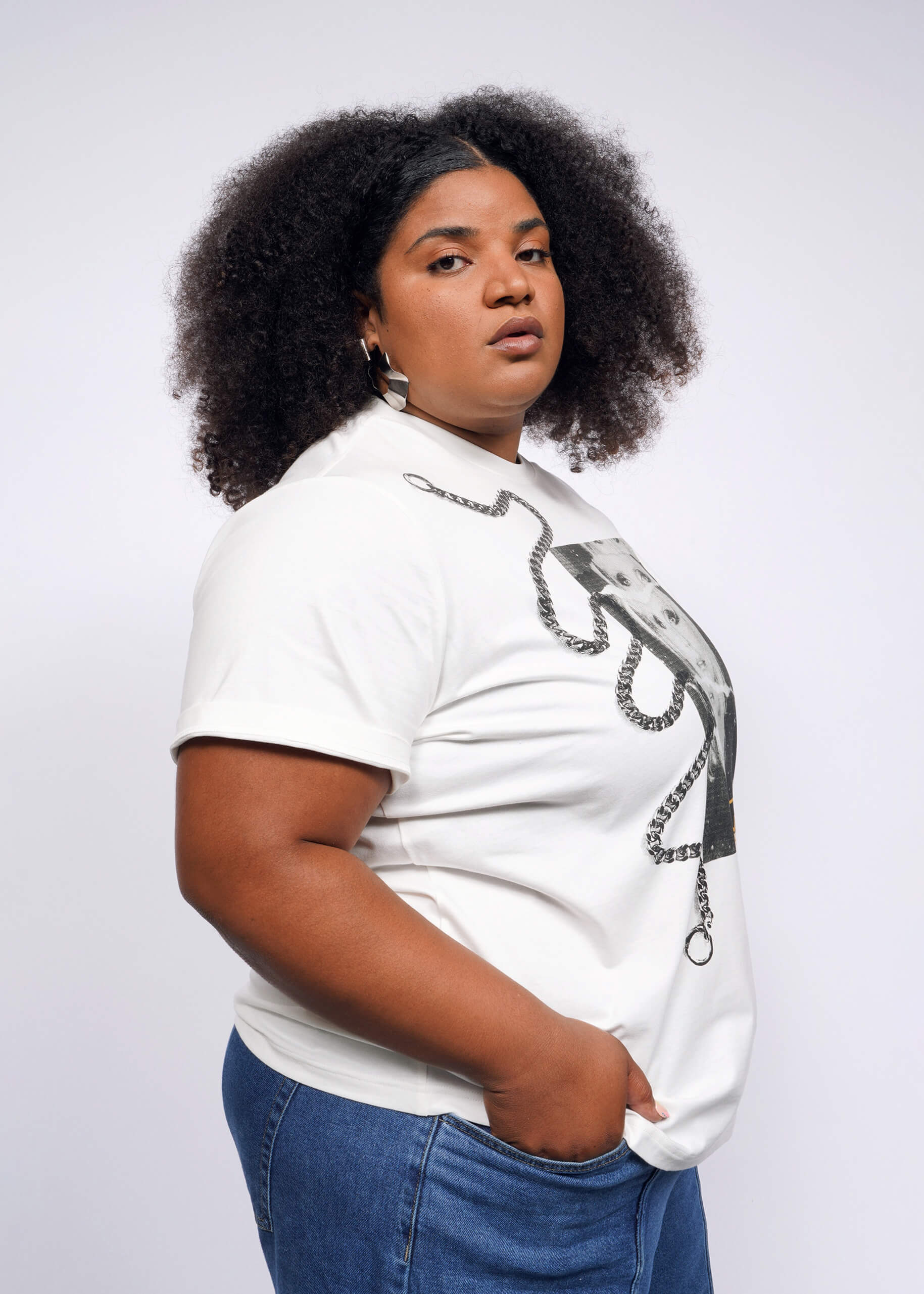 A person with curly hair channels a Debbie Harry vibe, wearing the Debbie Harry X Wildfang Graphic Tee and blue jeans. They stand confidently against a plain background, looking directly at the camera with one hand in their pocket, exuding a subtle punk aesthetic.