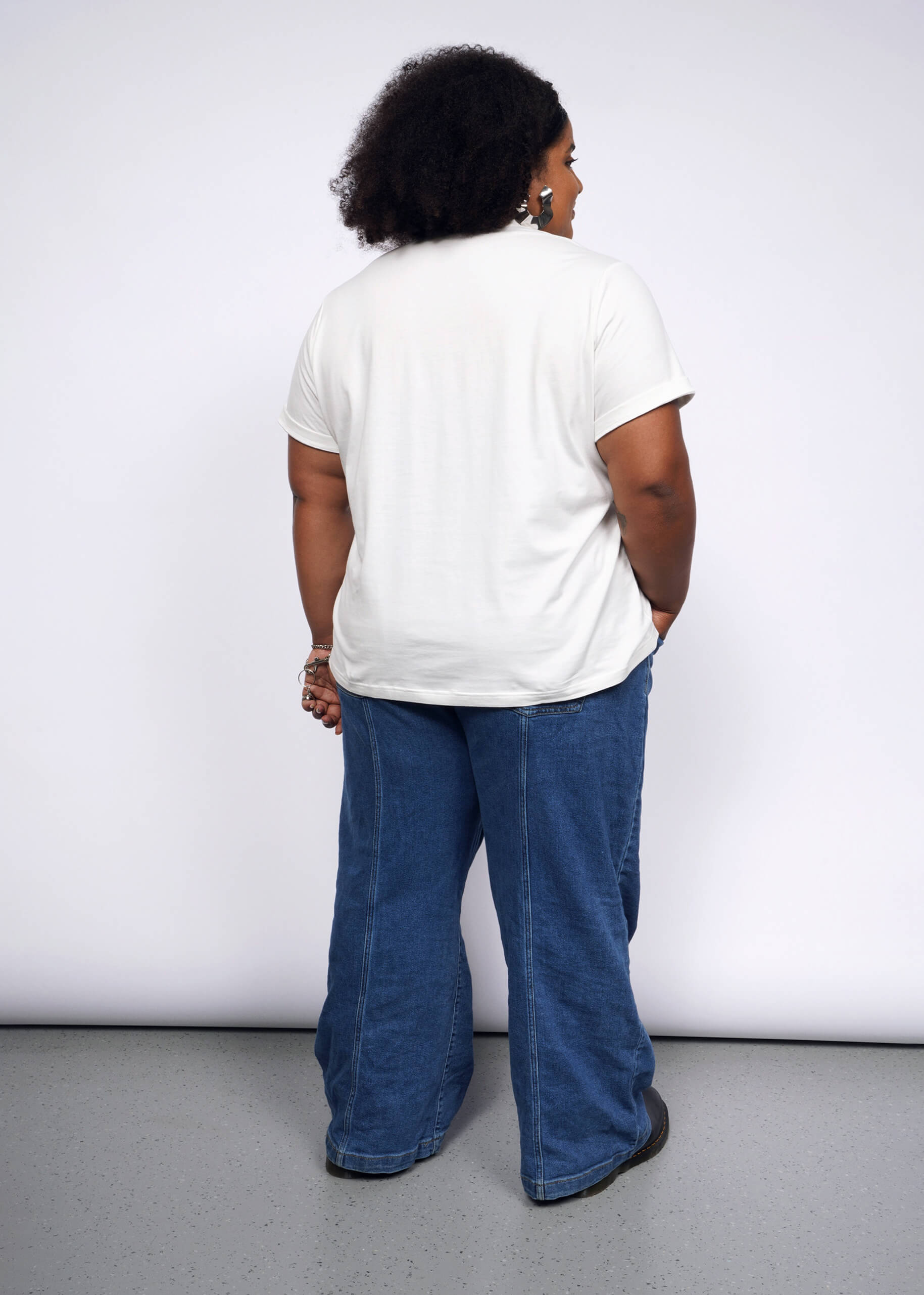 A person with curly hair faces away from the camera against a white backdrop, embracing a punk vibe. Theyre wearing the Debbie Harry X Wildfang Graphic Tee and wide-legged blue jeans, their hands relaxed by their sides.