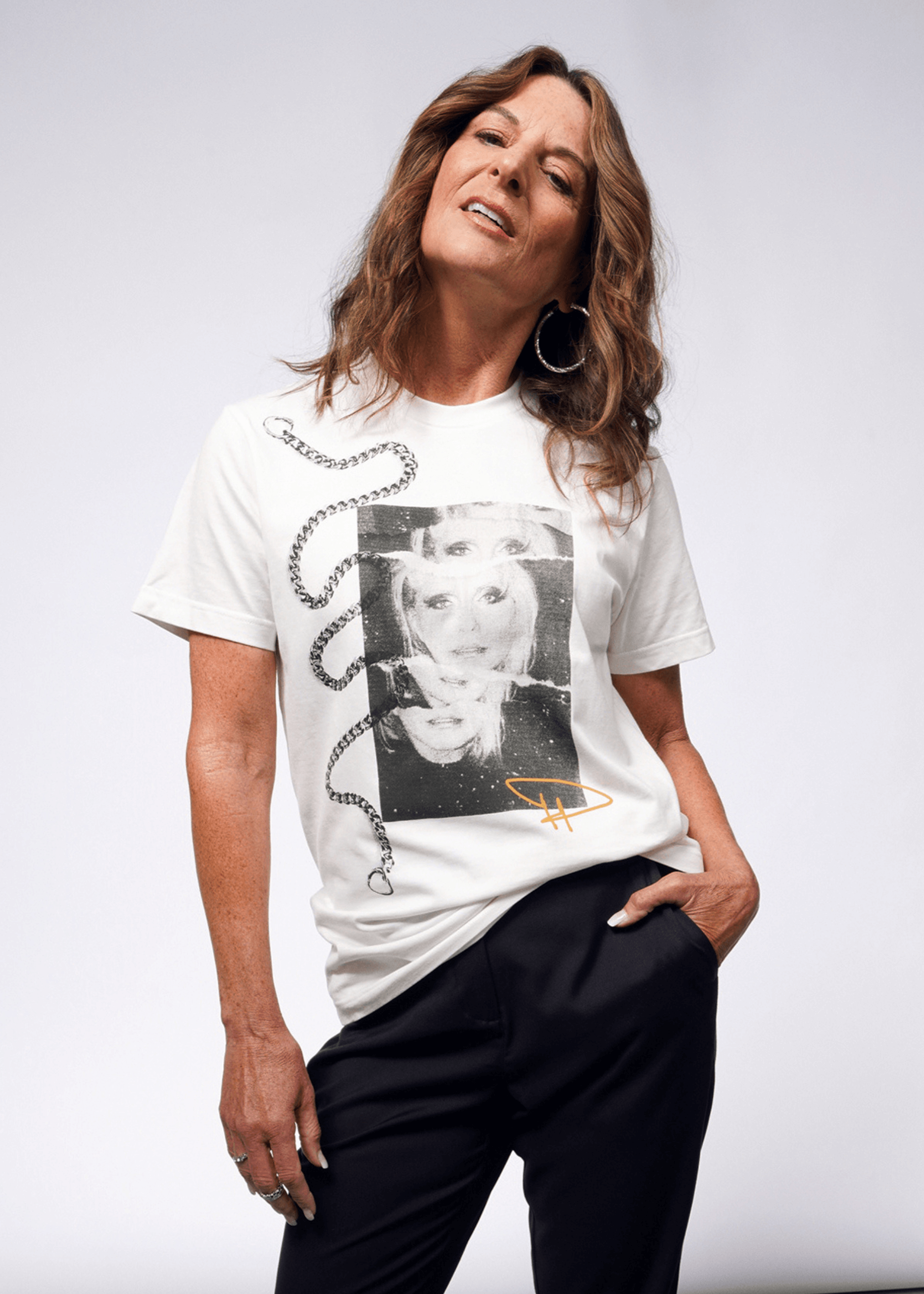 A person with long hair strikes a confident pose in the Debbie Harry X Wildfang Graphic Tee, showcasing an artistic black-and-white graphic and chain design for a punk vibe. Paired with dark pants, they look upward, hand in pocket, against a plain backdrop.