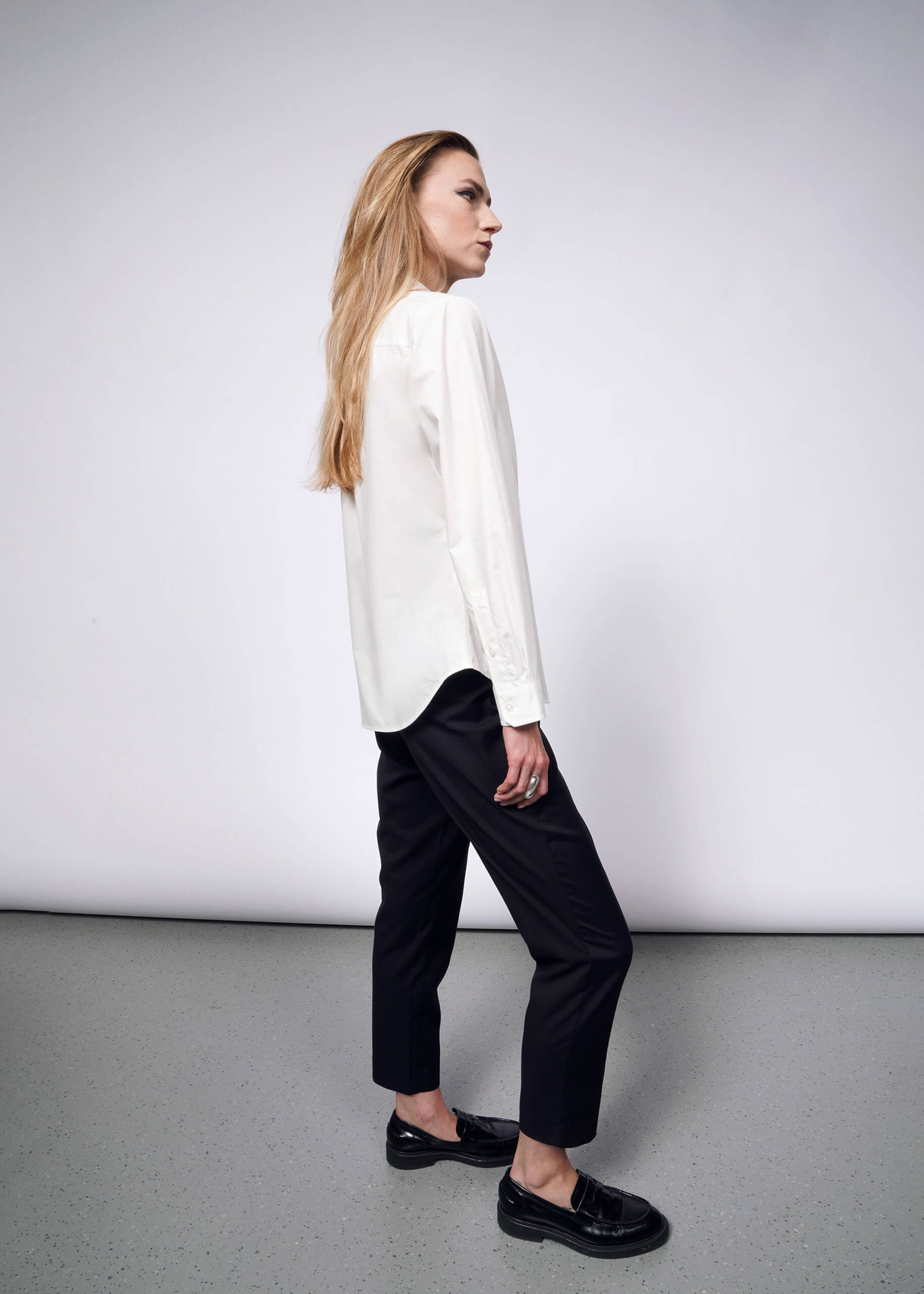 Against a plain backdrop, a person with long hair stands in profile wearing the Debbie Harry X Wildfang Long Sleeve Button Up featuring removable chain detail, paired with dark pants and black shoes. Channeling a punk aesthetic, their neutral expression is complemented by hands resting by their sides.