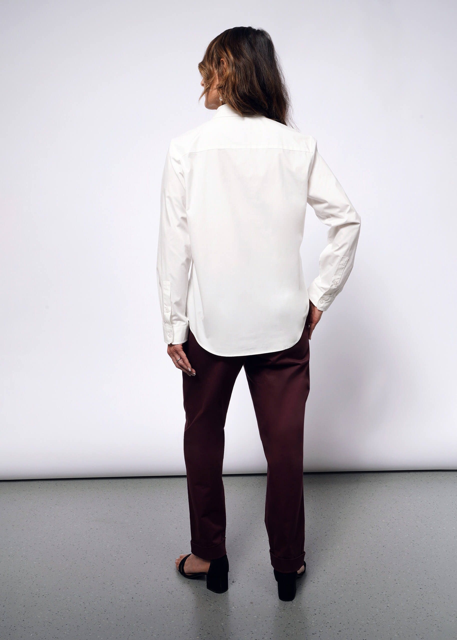 A person with long hair stands facing a white wall in a subtle punk style, wearing the Debbie Harry X Wildfang Long Sleeve Button Up, maroon pants with removable chain details, and black open-toe sandals. Their confident pose contrasts the gray floor beneath.