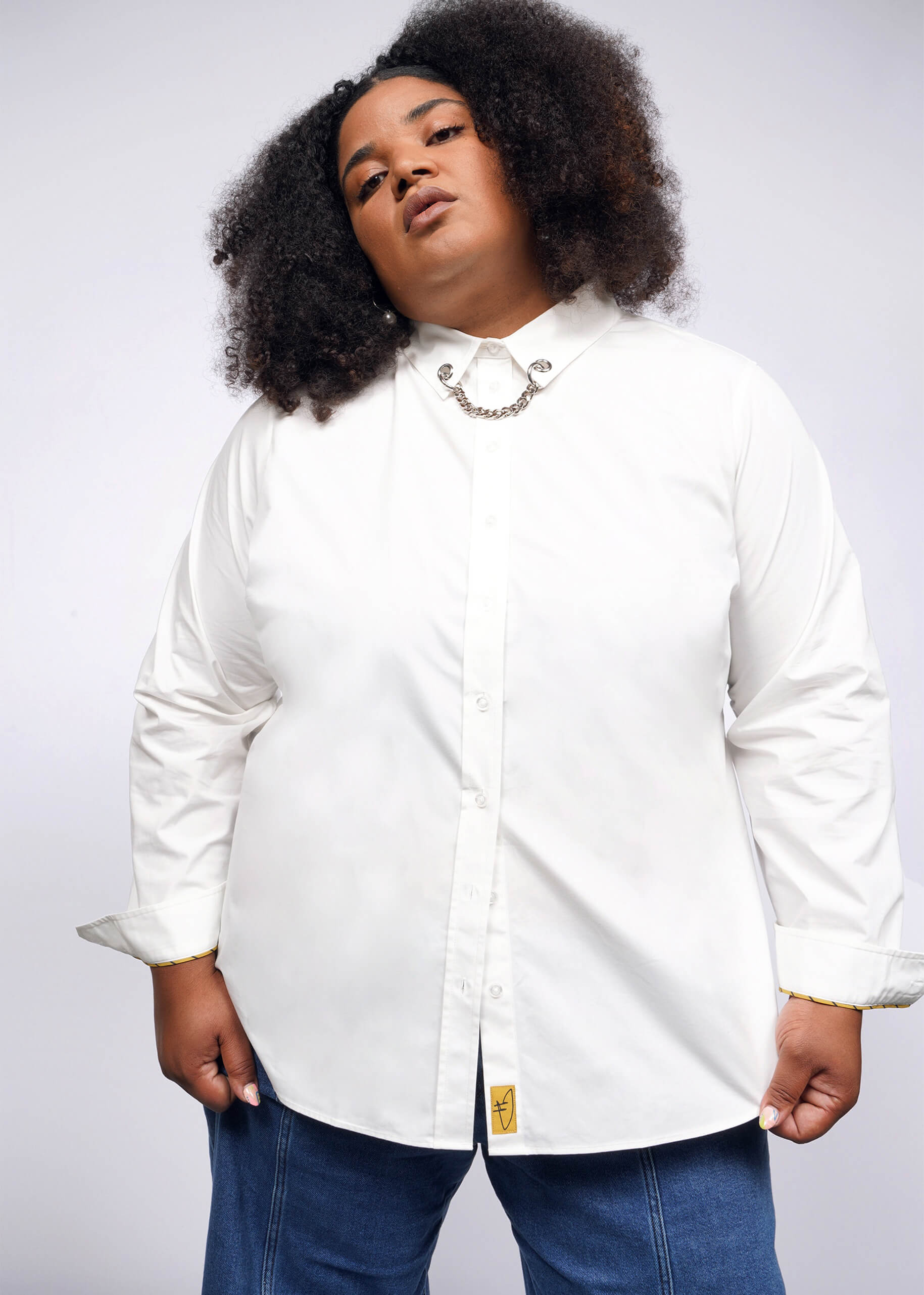 A person with curly hair confidently gazes at the camera, wearing a Debbie Harry X Wildfang Long Sleeve Button Up in white. It features a removable chain detail near the collar and a small yellow tag at the hem, adding a punk aesthetic to their blue jeans outfit.