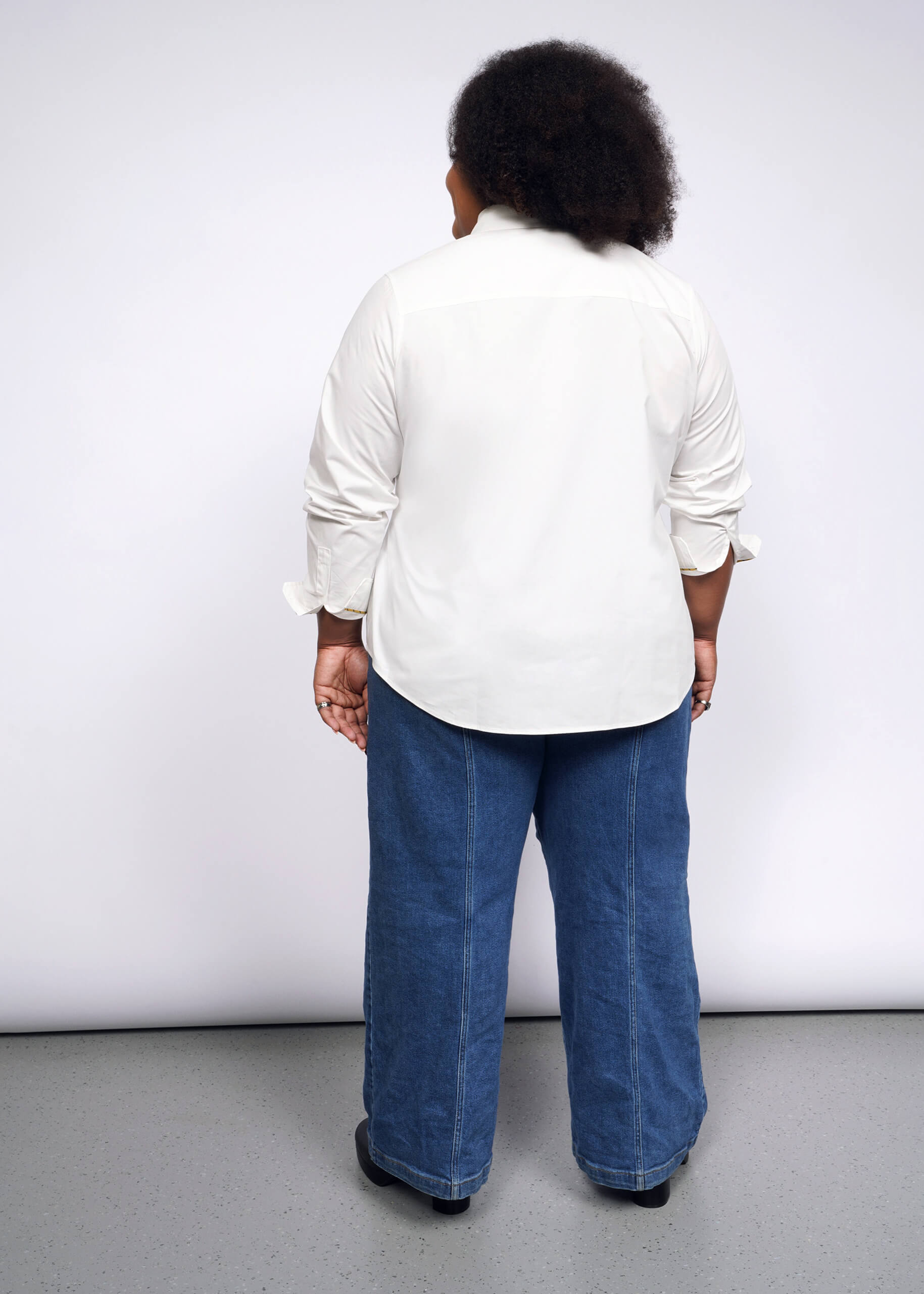 A person with a punk aesthetic and short curly hair stands with their back to the camera wearing the Debbie Harry X Wildfang Long Sleeve Button Up and blue jeans. Their slightly rolled-up sleeves enhance their cool vibe against a grey floor and plain white background.