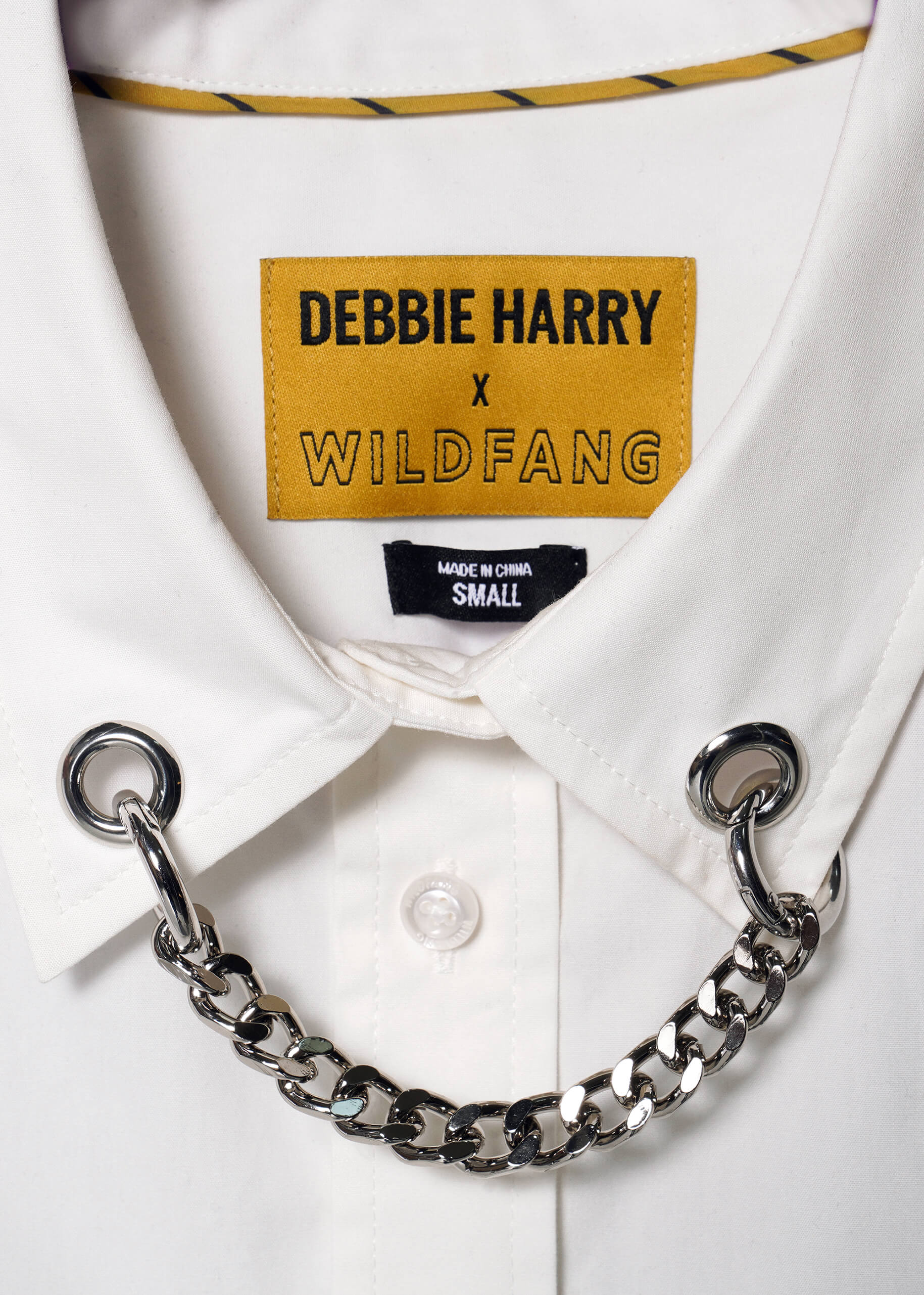 A close-up of a white shirt collar with metal eyelets and a shiny silver chain captures the punk aesthetic. Above it reads DEBBIE HARRY X WILDFANG, with MADE IN CHINA SMALL printed beneath, featuring a removable chain detail in the Debbie Harry X Wildfang Long Sleeve Button Up.