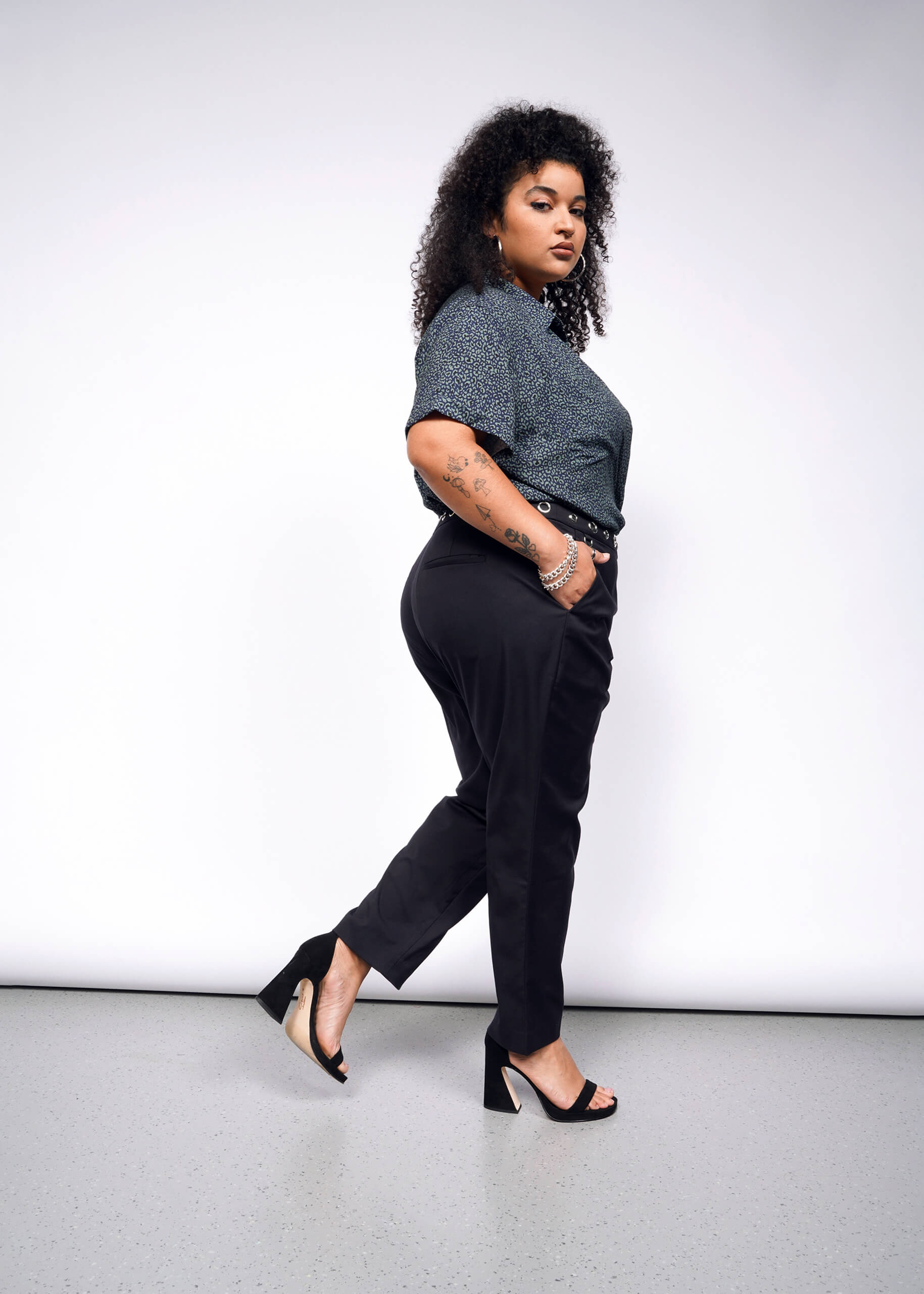 With curly hair, a person channels a punk vibe, confidently posing in a dark speckled top, Debbie Harry X Wildfang Slim Crop Pant, and high heels. The plain white background highlights their bold look as they stride forward.