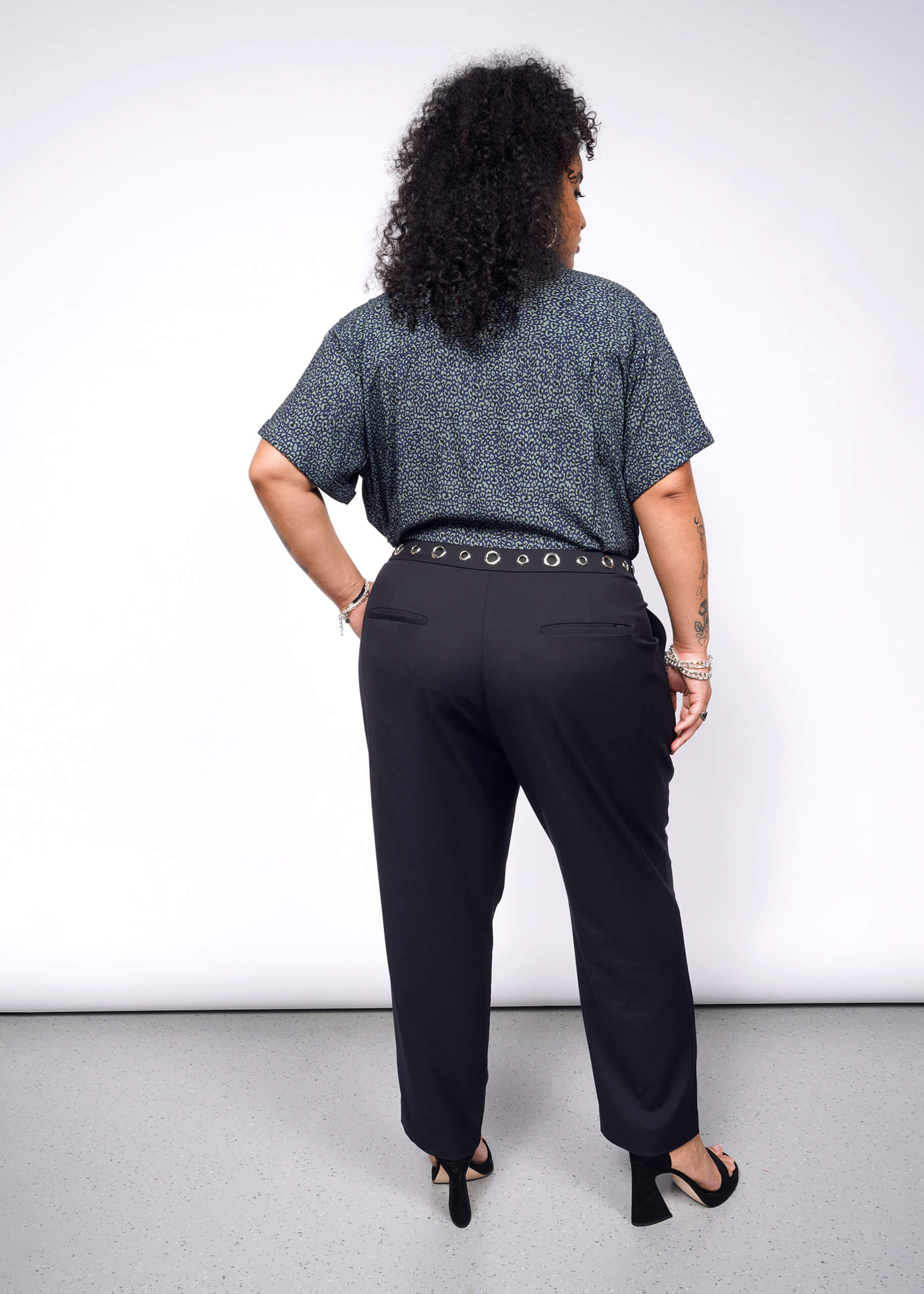 With curly hair exuding a subtle punk vibe, they stand out in a blue-patterned Wildfang collection shirt and Debbie Harry X Wildfang Slim Crop Pant. Black heels and bracelets on their wrist add an edgy flair against the plain background.