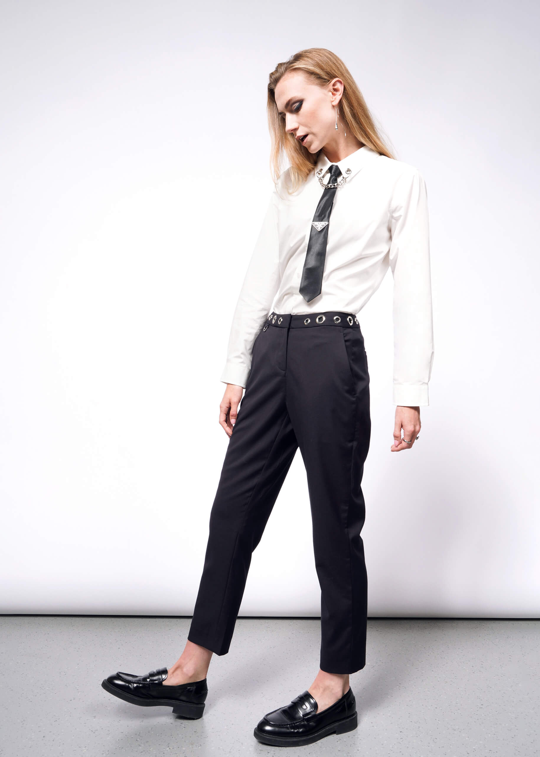 A person with long hair channels a punk aesthetic akin to Debbie Harry, standing relaxed against a plain backdrop. They wear a white shirt, black tie, and black loafers from the Wildfang collection, paired with the Debbie Harry X Wildfang Slim Crop Pant. Their head is slightly tilted downwards.