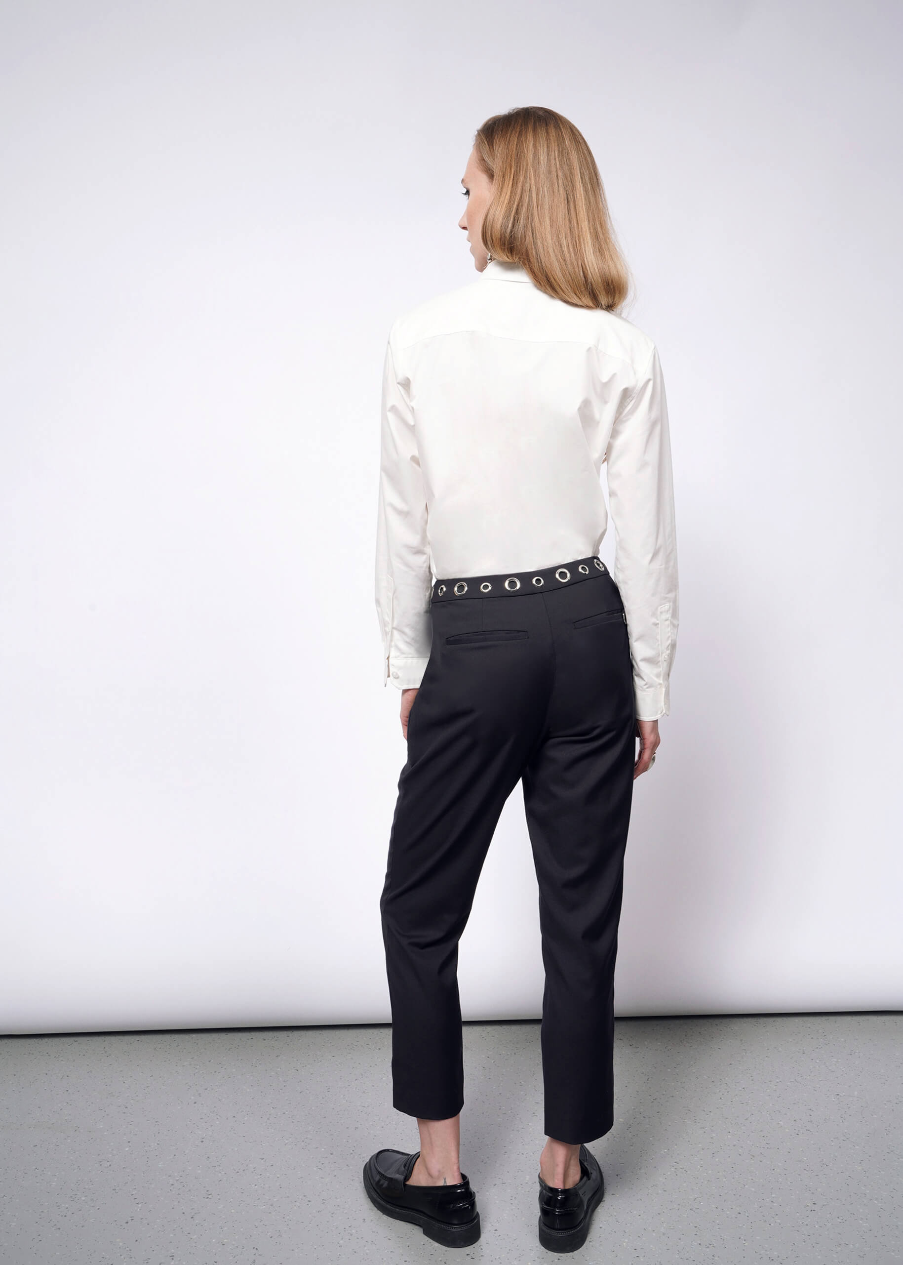 A person with long hair, in a white button-up shirt and Debbie Harry X Wildfang Slim Crop Pants featuring a studded belt, stands facing away in a minimalist setting. The gray floor and plain white wall complete the punk-inspired aesthetic of the Wildfang collection.