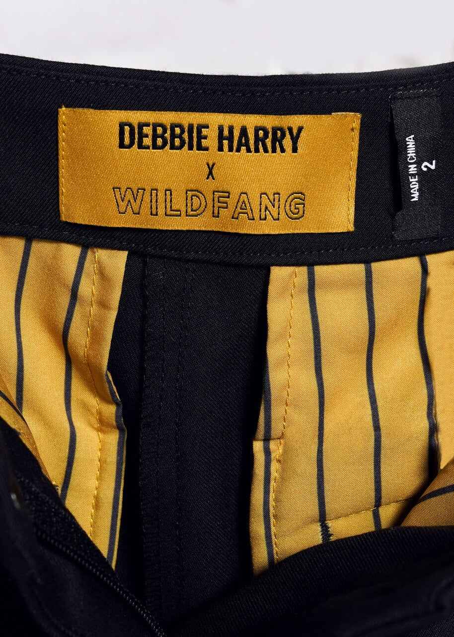 Close-up of a clothing label from the Debbie Harry X Wildfang Slim Crop Pant with a punk aesthetic: black text on yellow, yellow and black striped lining, and a bold black size tag with white text on the right.