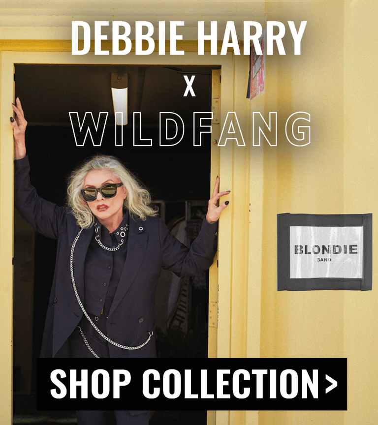 Debbie Harry stands in doorway of yellow metal building with the sign BLONDIE on the outside. She is wearing sunglasses and pieces from the Debbie Harry x Wildfang collection. Text overlay says DEBBIE HARRY X WILDFANG. Button on bottom of image says SHOP COLLECTION.