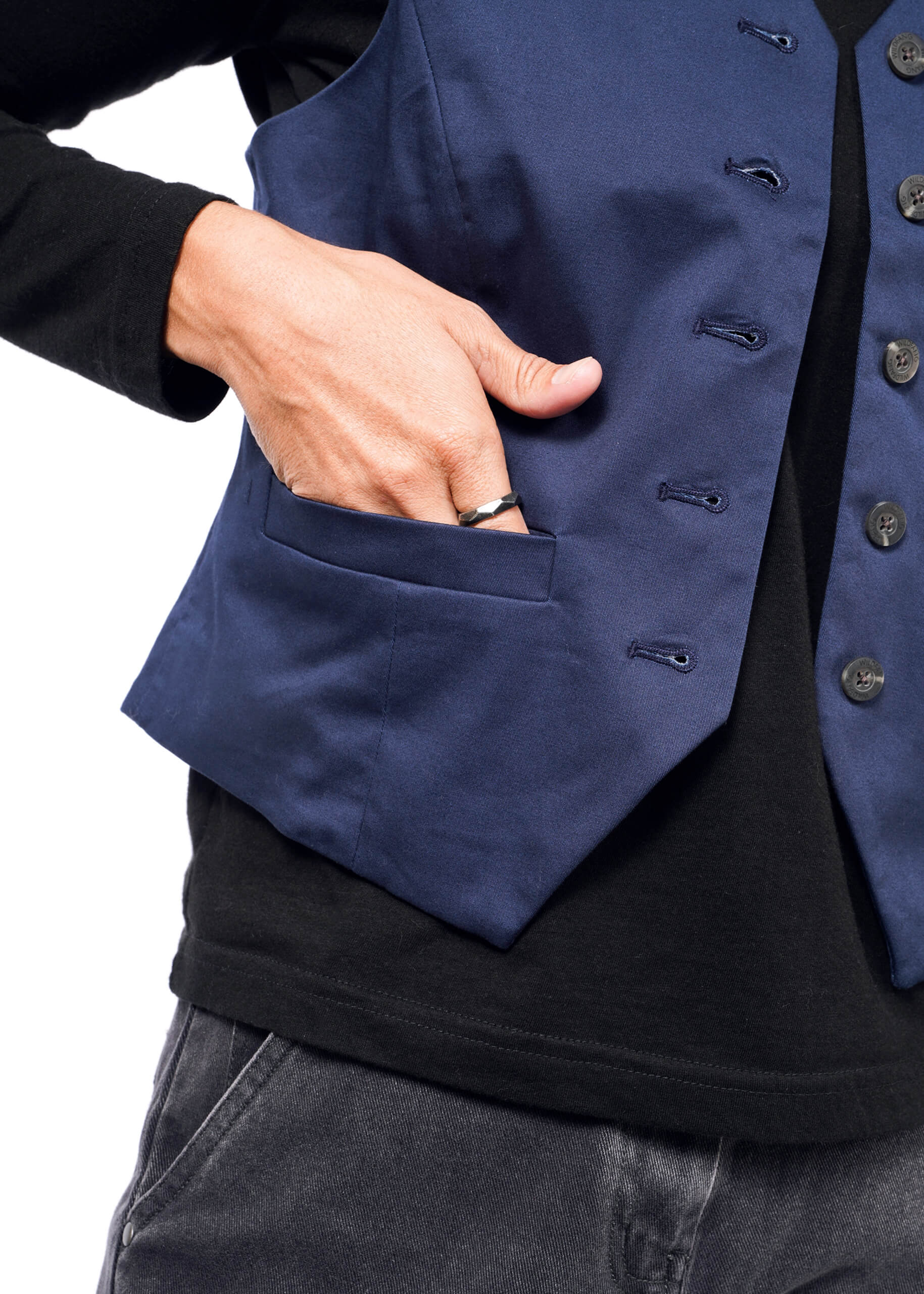 Wearing The Essential Vest in tailored fit dark blue over a black shirt, a person places their hand in its pocket. The stretch cotton, ideal for layering, has front buttons. A ring adorns their thumb.