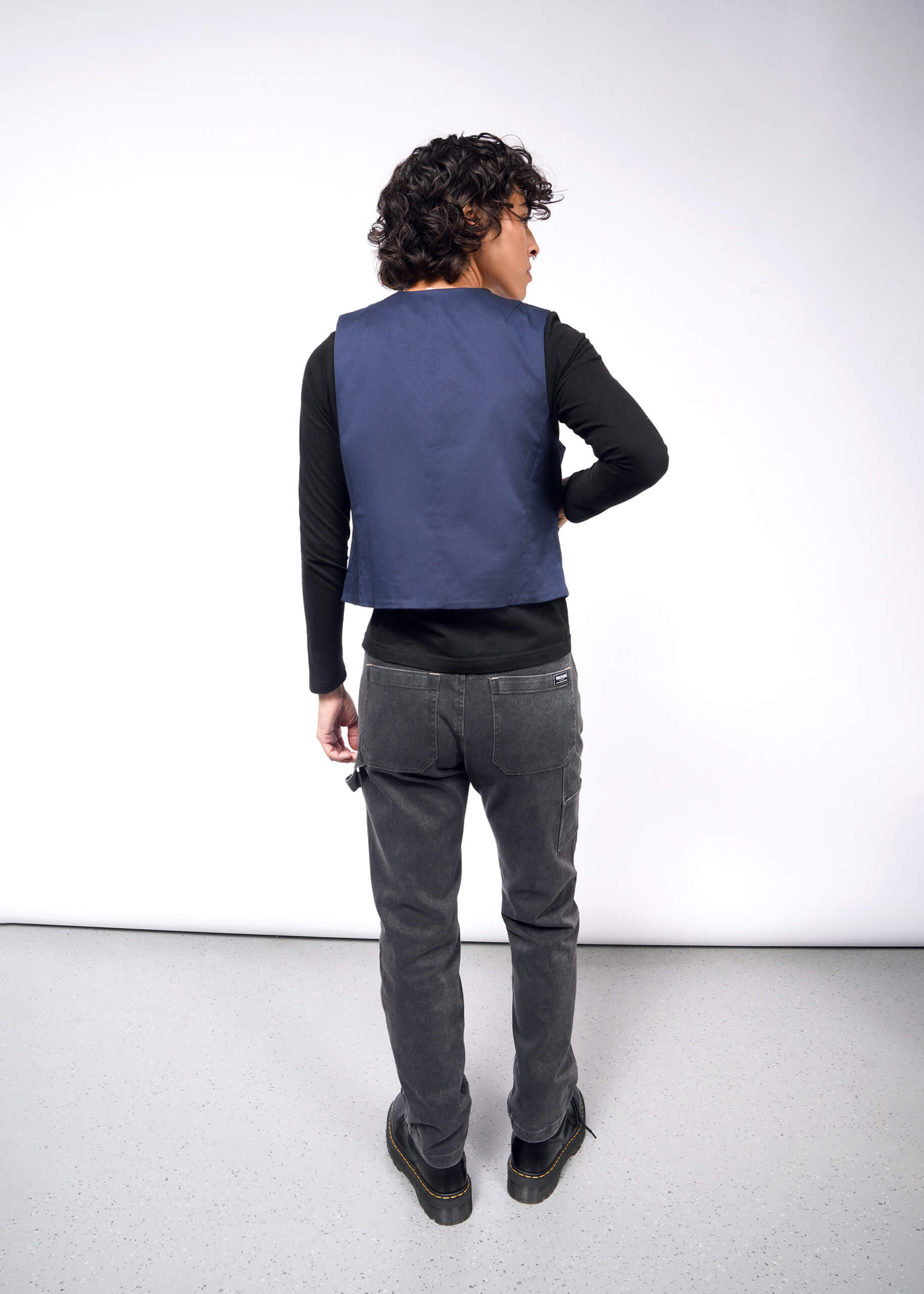 A man with curly hair is wearing a black long-sleeve shirt, The Essential Vest tailored fit in stretch cotton, and gray jeans, standing on a light gray floor facing a white wall.