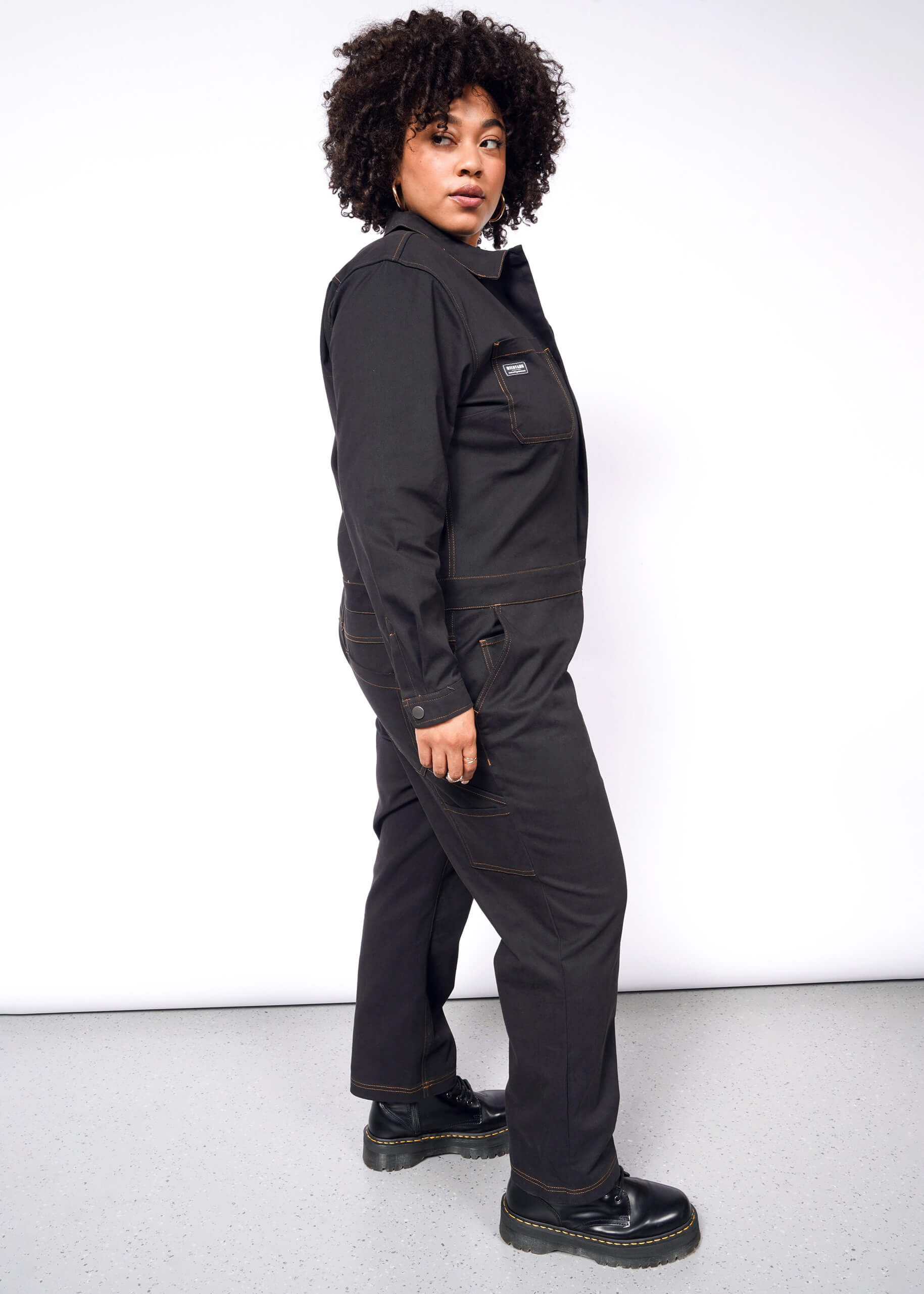 A person with curly hair stands sideways, gazing at the camera in a black Essential Long Sleeve Coverall made from brushed stretch cotton. It has an oversized fit and 10 pockets, paired with sleek black boots against a plain white background.