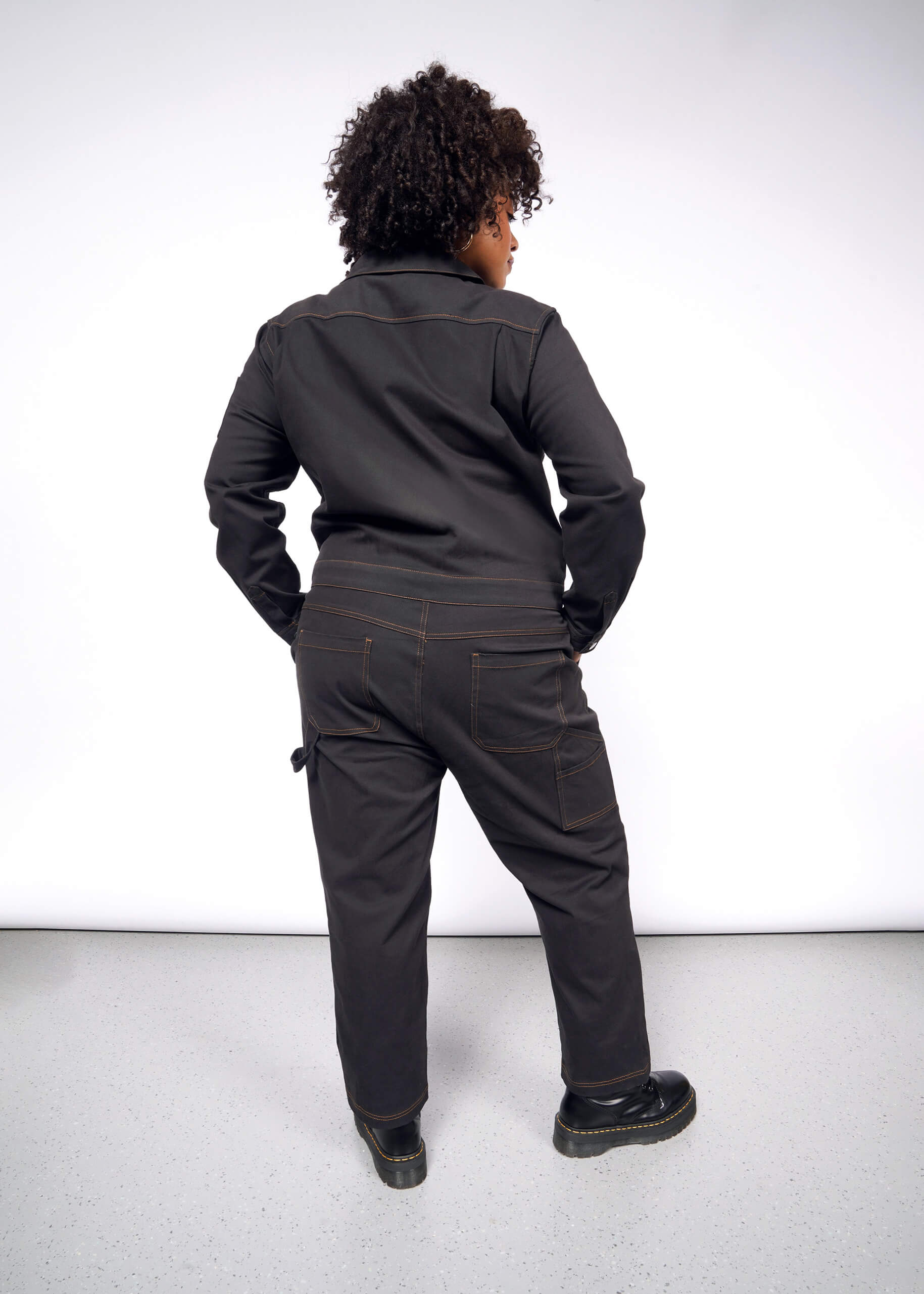 A person with curly hair faces away from the camera, wearing The Essential Long Sleeve Coverall, an oversized black jumpsuit crafted in brushed stretch cotton. They match it with black shoes atop a light gray floor, all set against a plain white background, exuding effortless style and function.