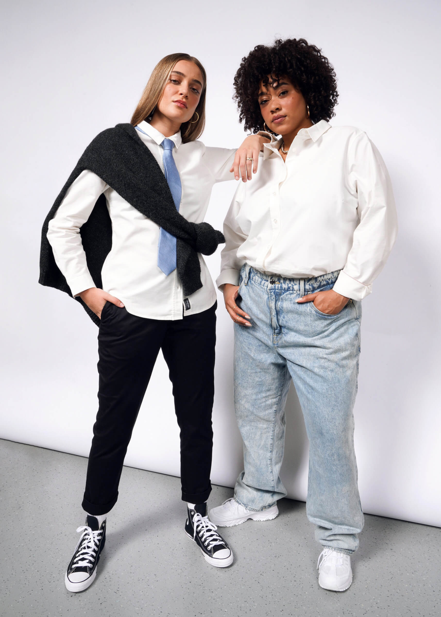 Two individuals pose confidently against a white background. One sports a tailored fit with The Essential Long Sleeve Oxford Button Up, black pants, blue tie, and black Converse sneakers. The other wears a 100% cotton shirt and relaxed blue jeans. Both exude a casual, stylish vibe.