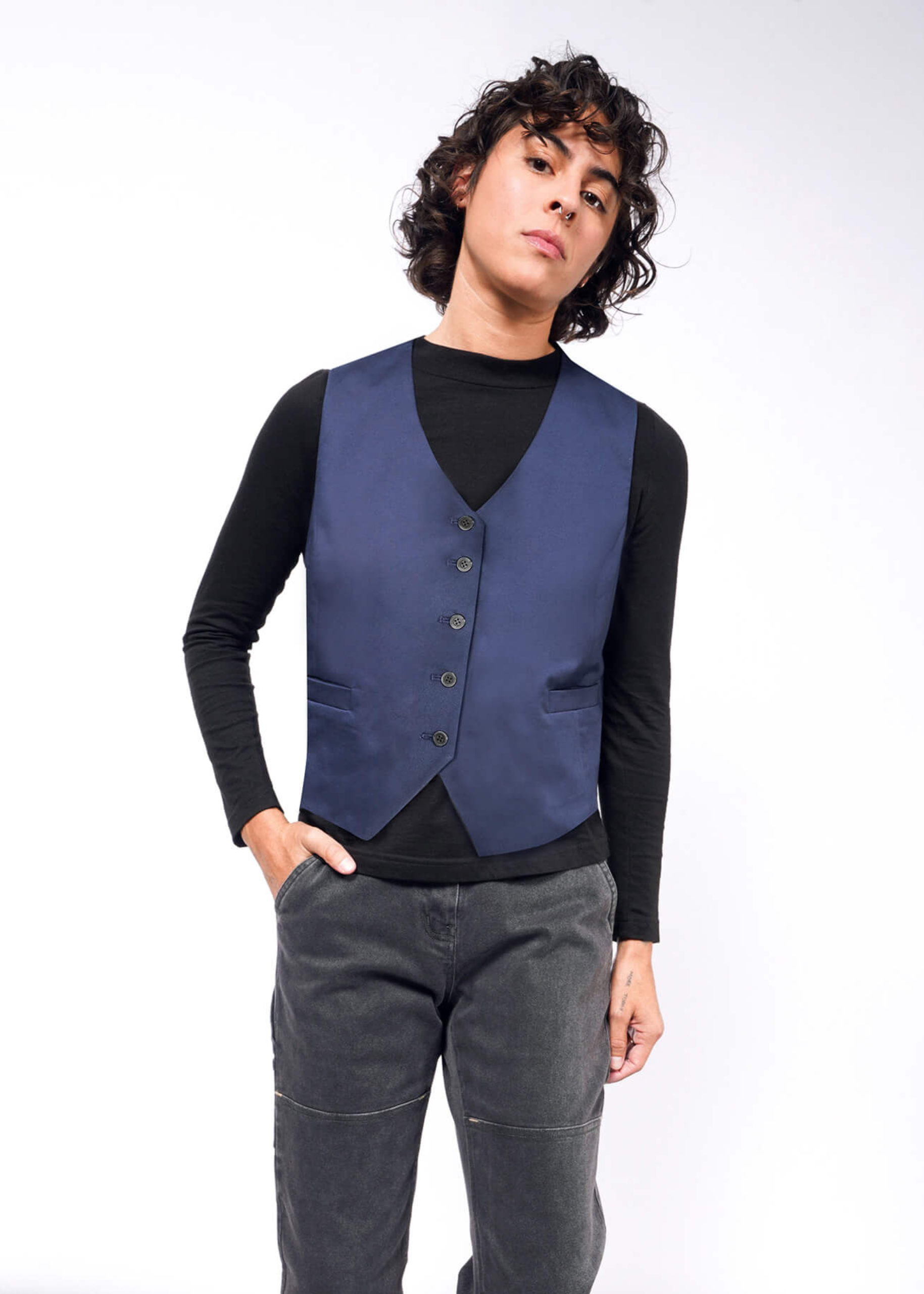 Modeling The Essential Vest in tailored fit blue stretch cotton over a black long-sleeve shirt and gray pants, a person with curly hair stands against a plain white background. With one hand in their pocket, they gaze into the camera neutrally, elegantly showcasing this layering piece.