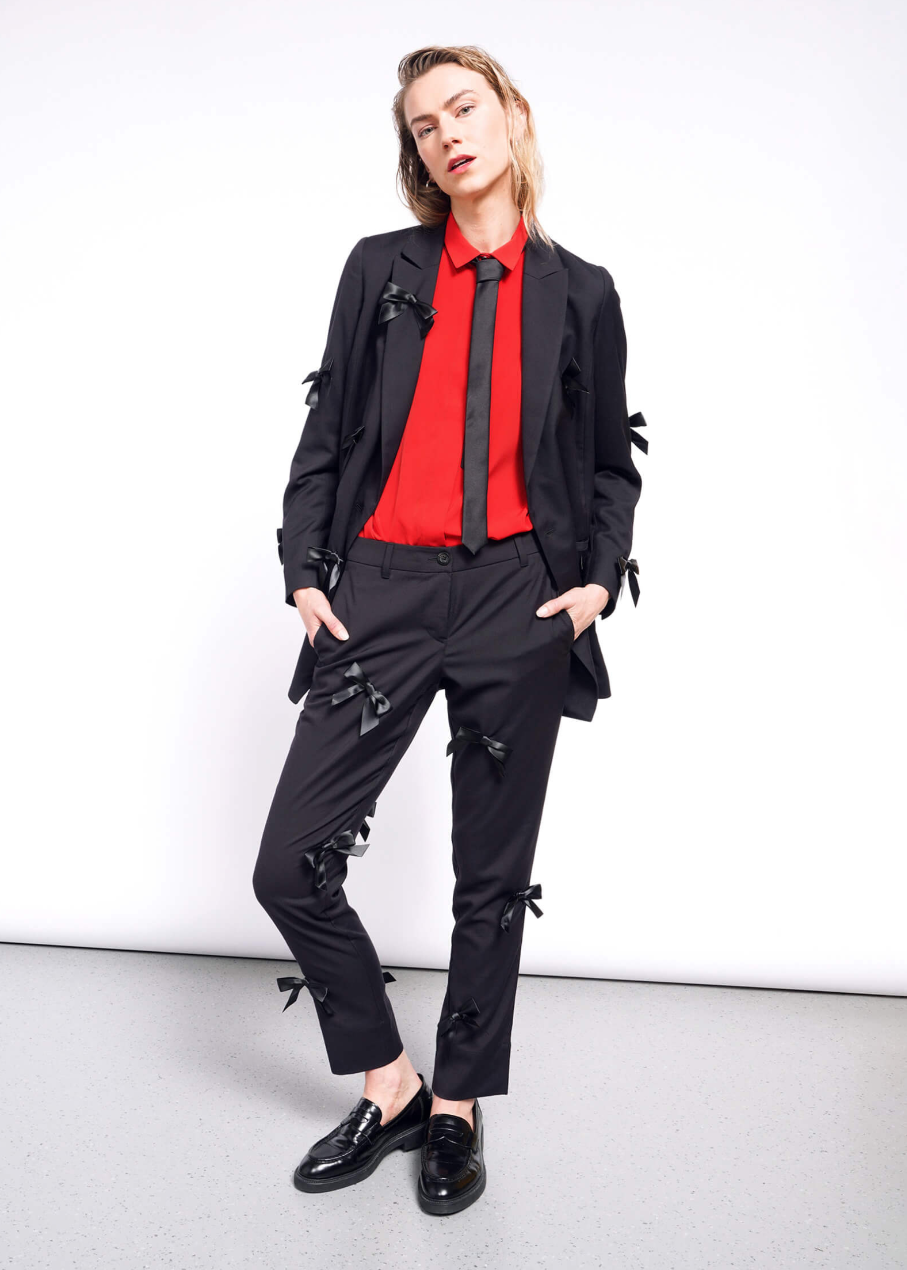 A person poses against a white background wearing a black suit with The Empower Bows Trouser, bow-embellished pants, a red shirt, black tie, and loafers. One hand rests in their pocket, exuding an ultra-luxe feel.