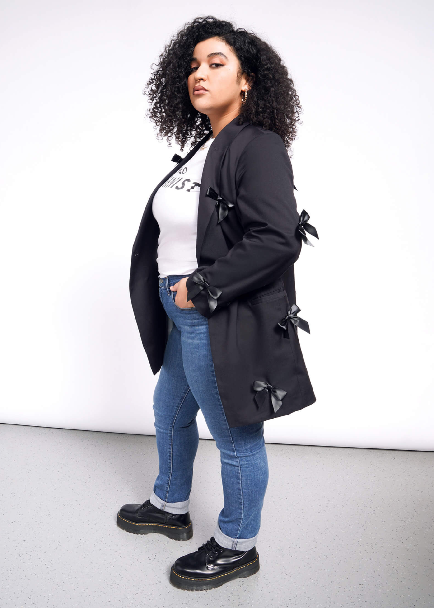 A person with curly hair stands confidently in a white t-shirt, blue jeans, and black shoes, wearing The Empower Bows Double Breasted Blazer adorned with decorative side bows. The background is a plain mix of white and gray.