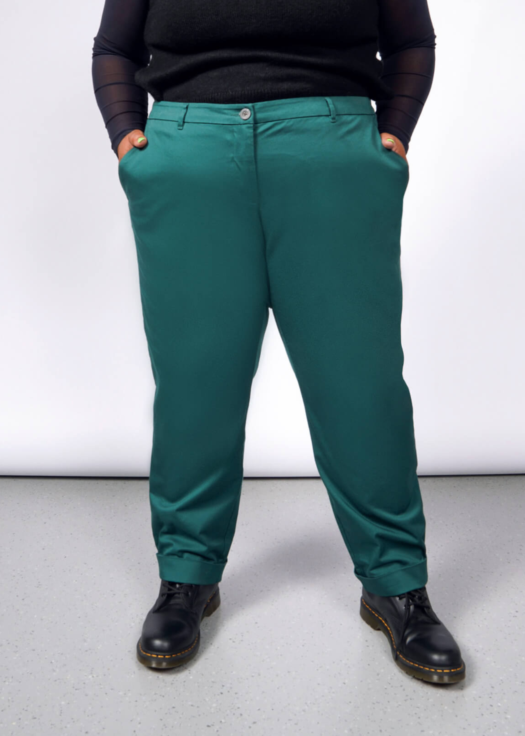 A person models The Essential Trouser in classic teal with black shoes, standing confidently with hands in pockets against a light gray backdrop, highlighting the timeless wardrobe staple.