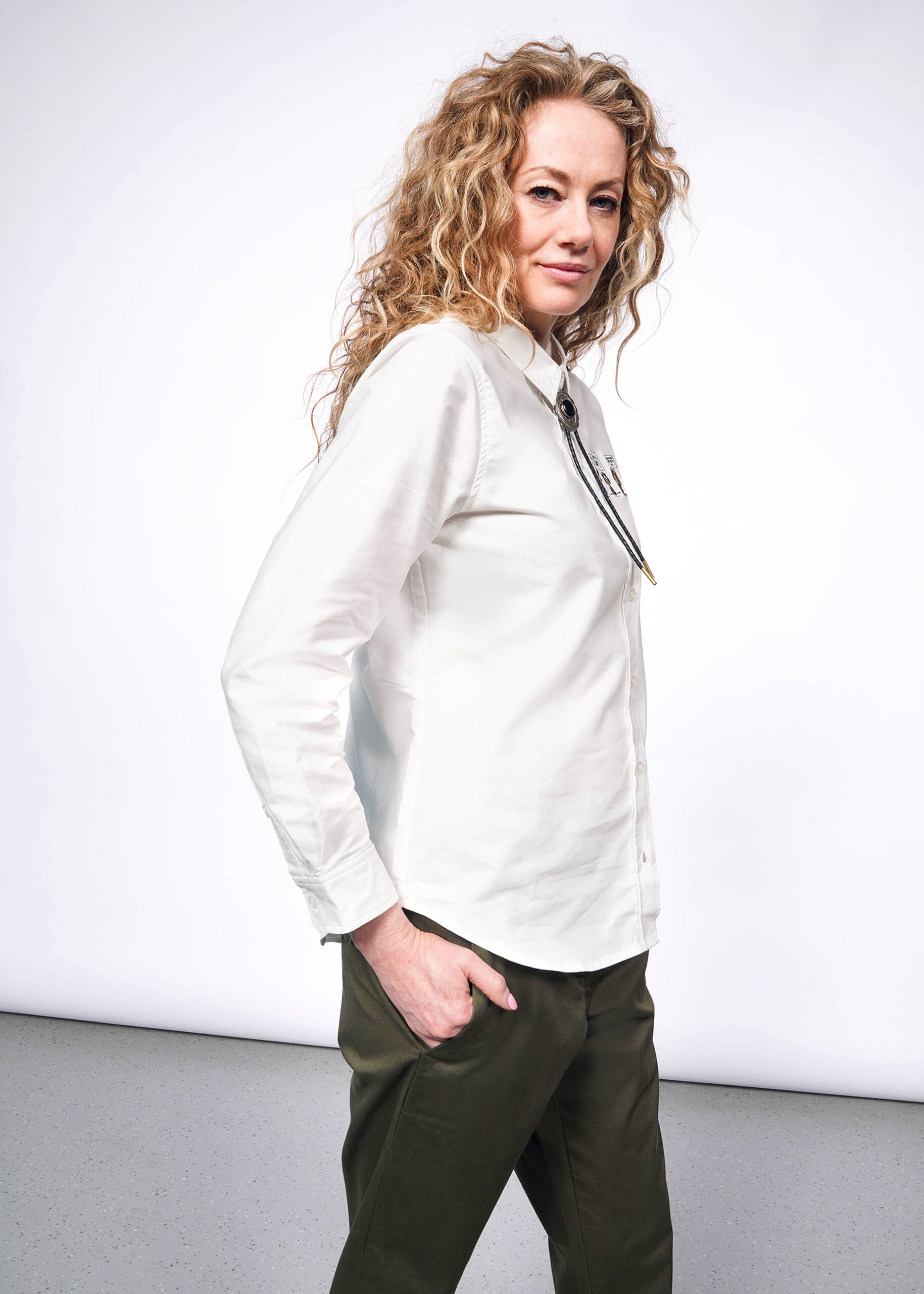 Side view of person with wavy blonde hair looking at camera wearing white long sleeve button up shirt with embroidered Peanuts characters, green trousers, and bolo tie.