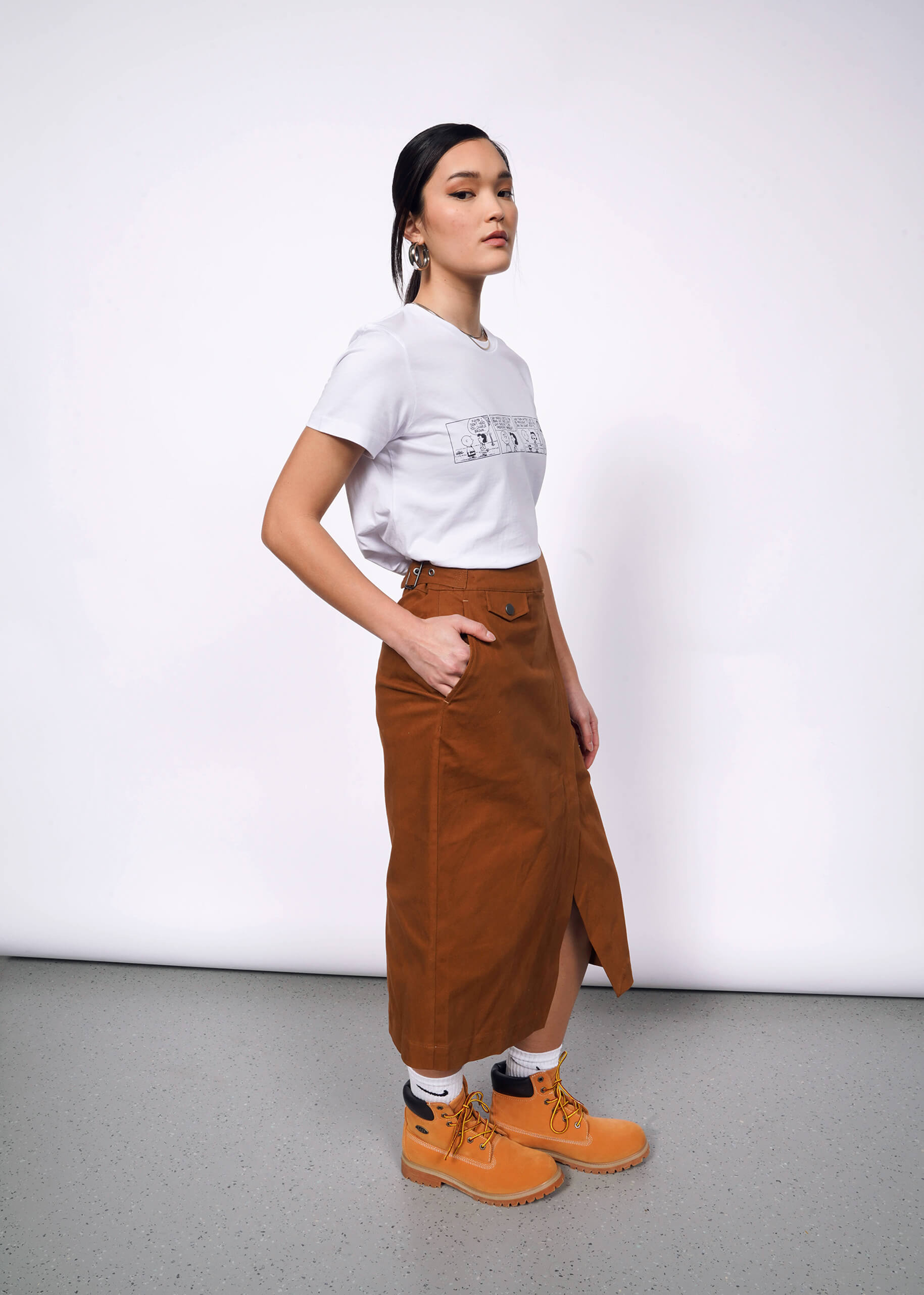 Side view of person with black hair tied back wearing white tshirt with Peanuts comic strip printed on it, brown skirt, and Timberland boots.